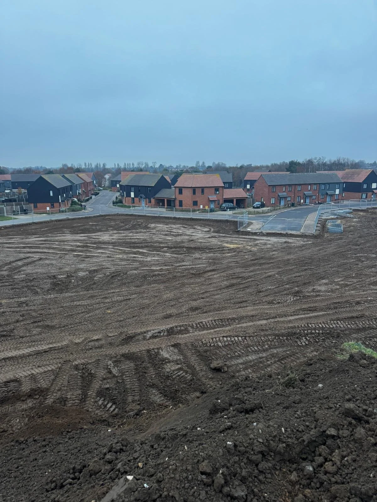 View of a construction site with freshly plowed dirt and tire tracks, residential houses in the background under a cloudy sky.