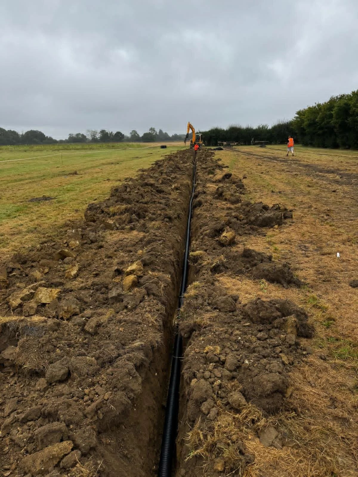 A long trench in the ground with a black pipe laid inside for drainage, with construction workers and machinery in the background under a cloudy sky.