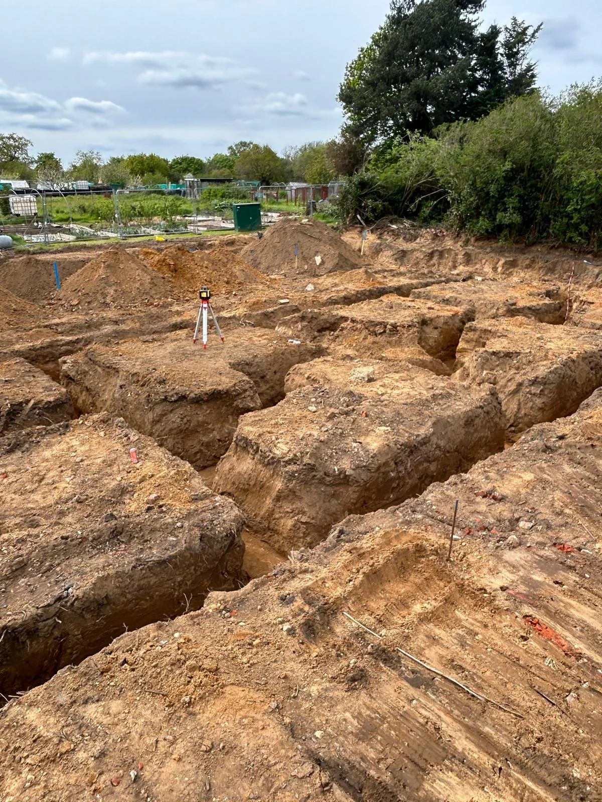 Underground excavation site with multiple trenches dug to form foundations and a laser level, surrounded by dirt mounds and trees in the background.