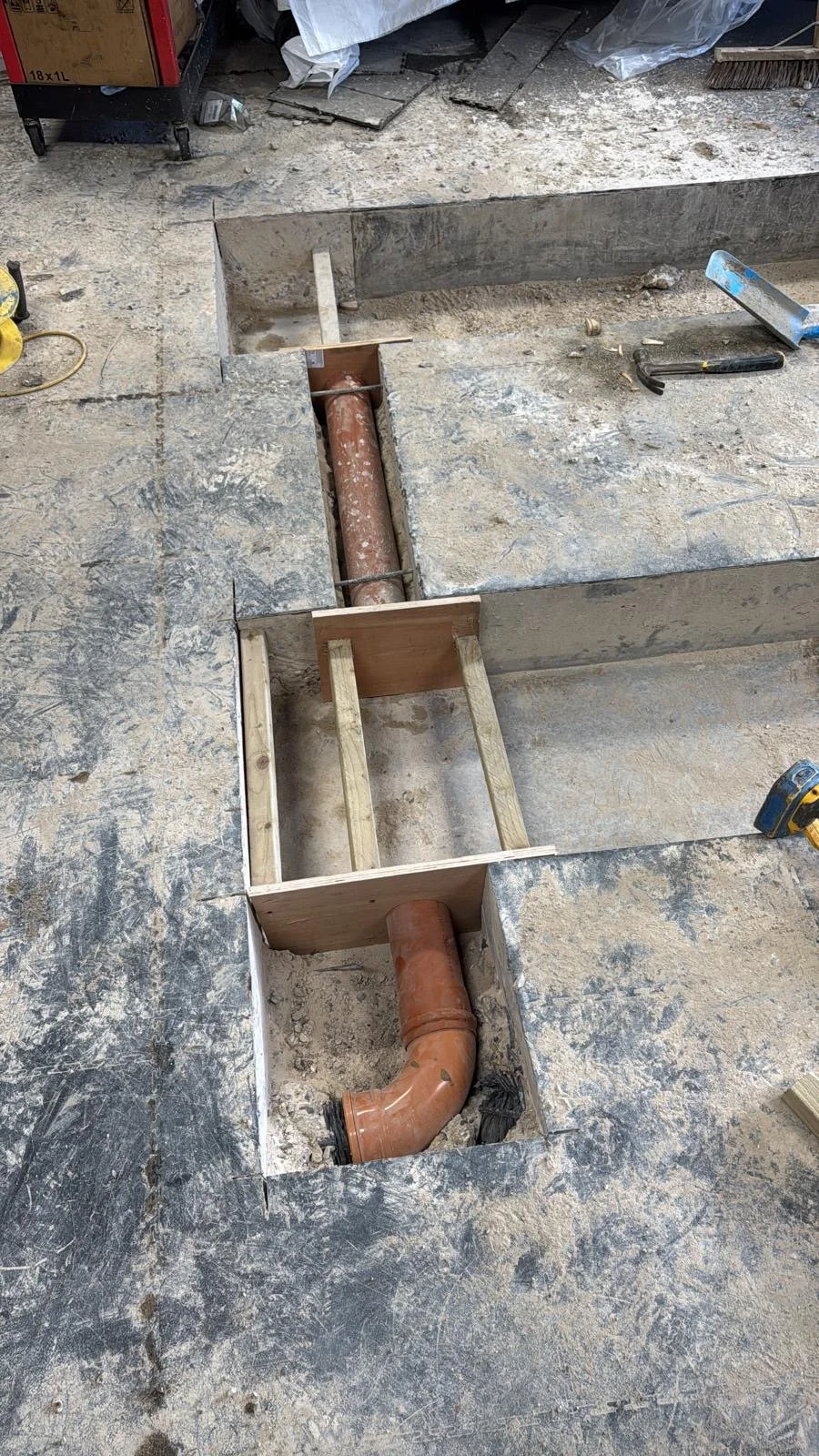 Automotive garage with open floor sections showing old and new pipes, wooden supports, and construction tools on the concrete floor.