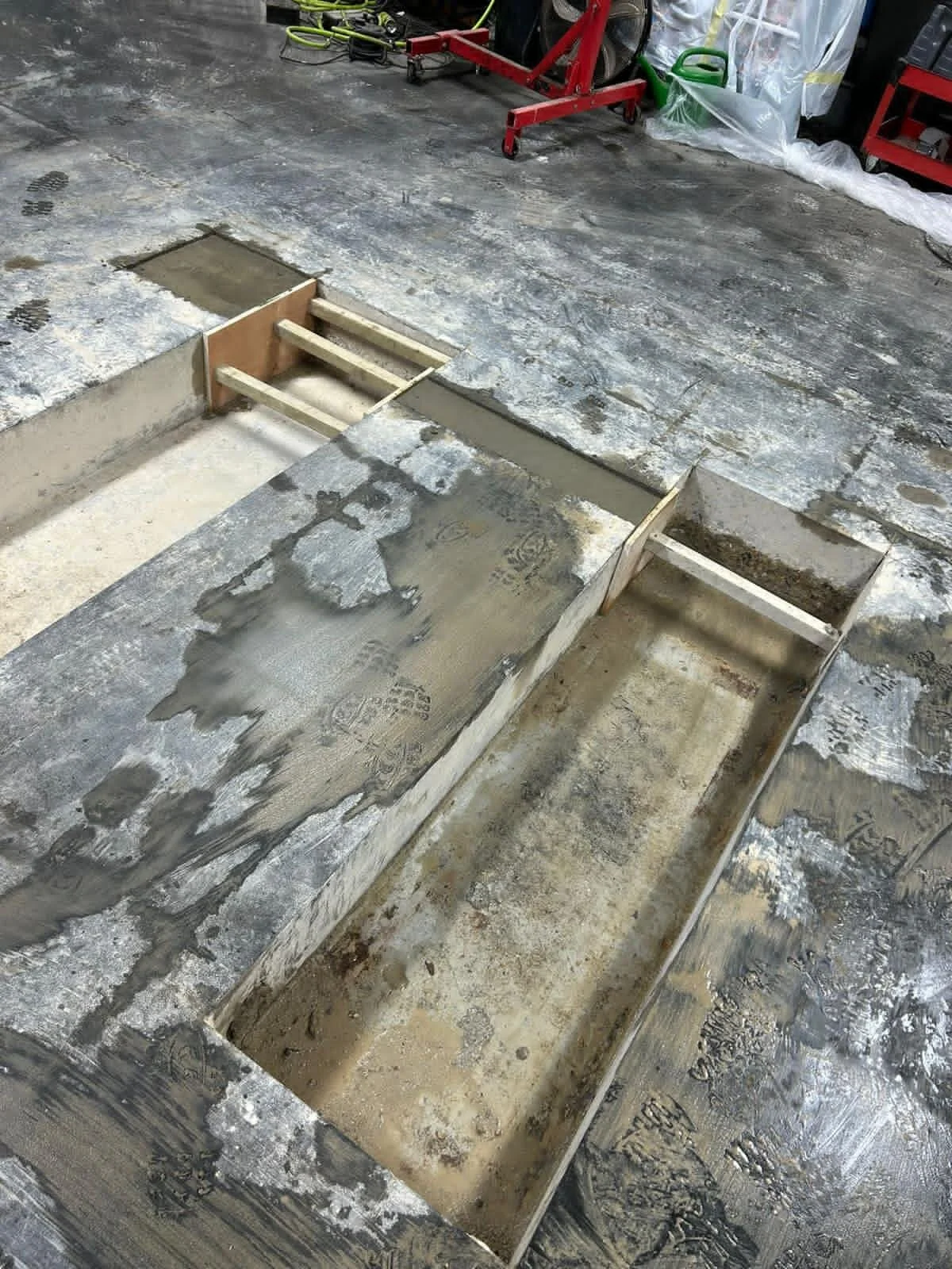 Automotive garage floor with wooden formwork for pouring concrete in a rectangular area, surrounded by a concrete floor with construction tools and materials in the background.