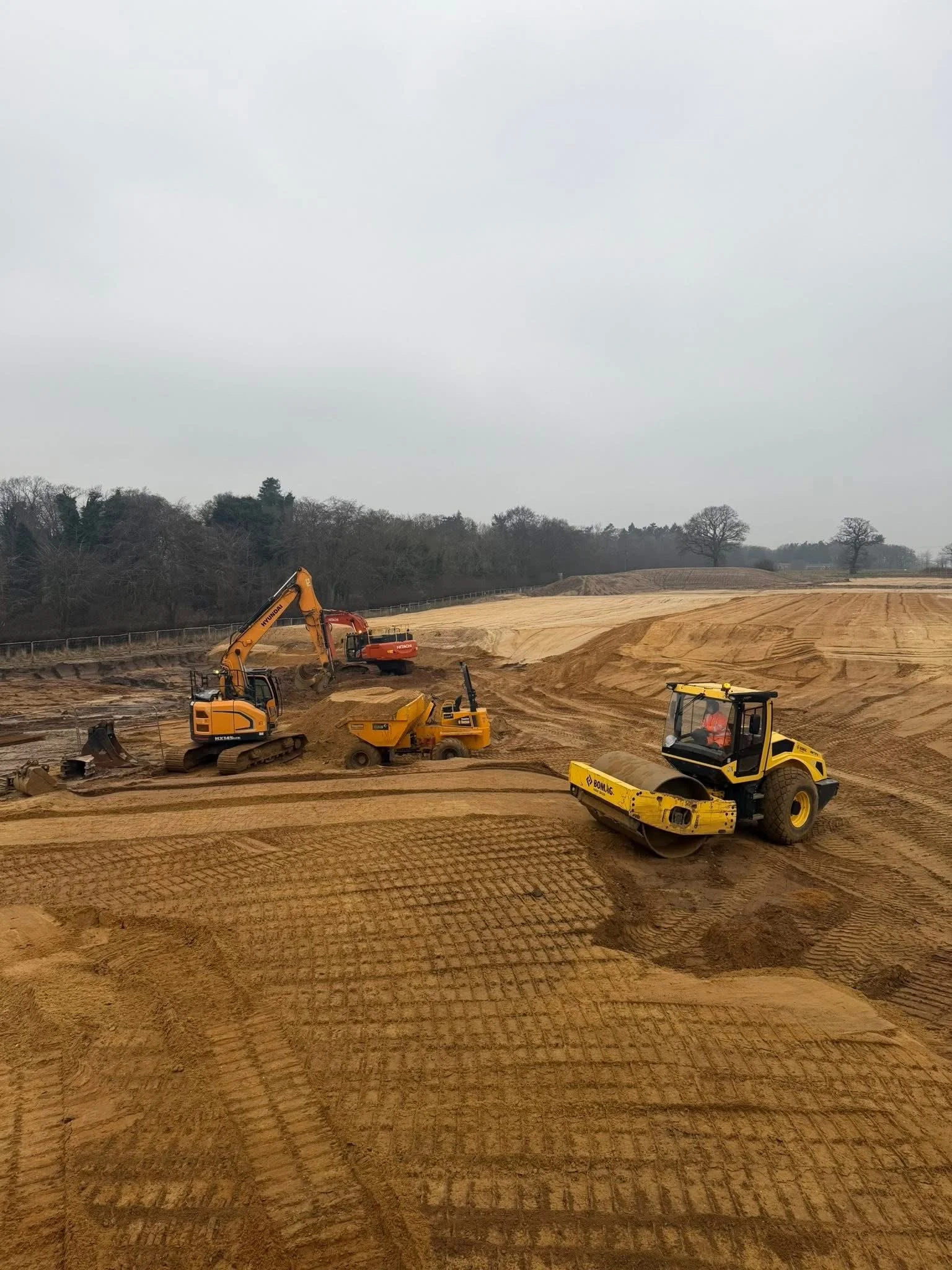 Construction site with yellow and orange excavators and a road roller working on a dirt road against a backdrop of trees and cloudy sky.