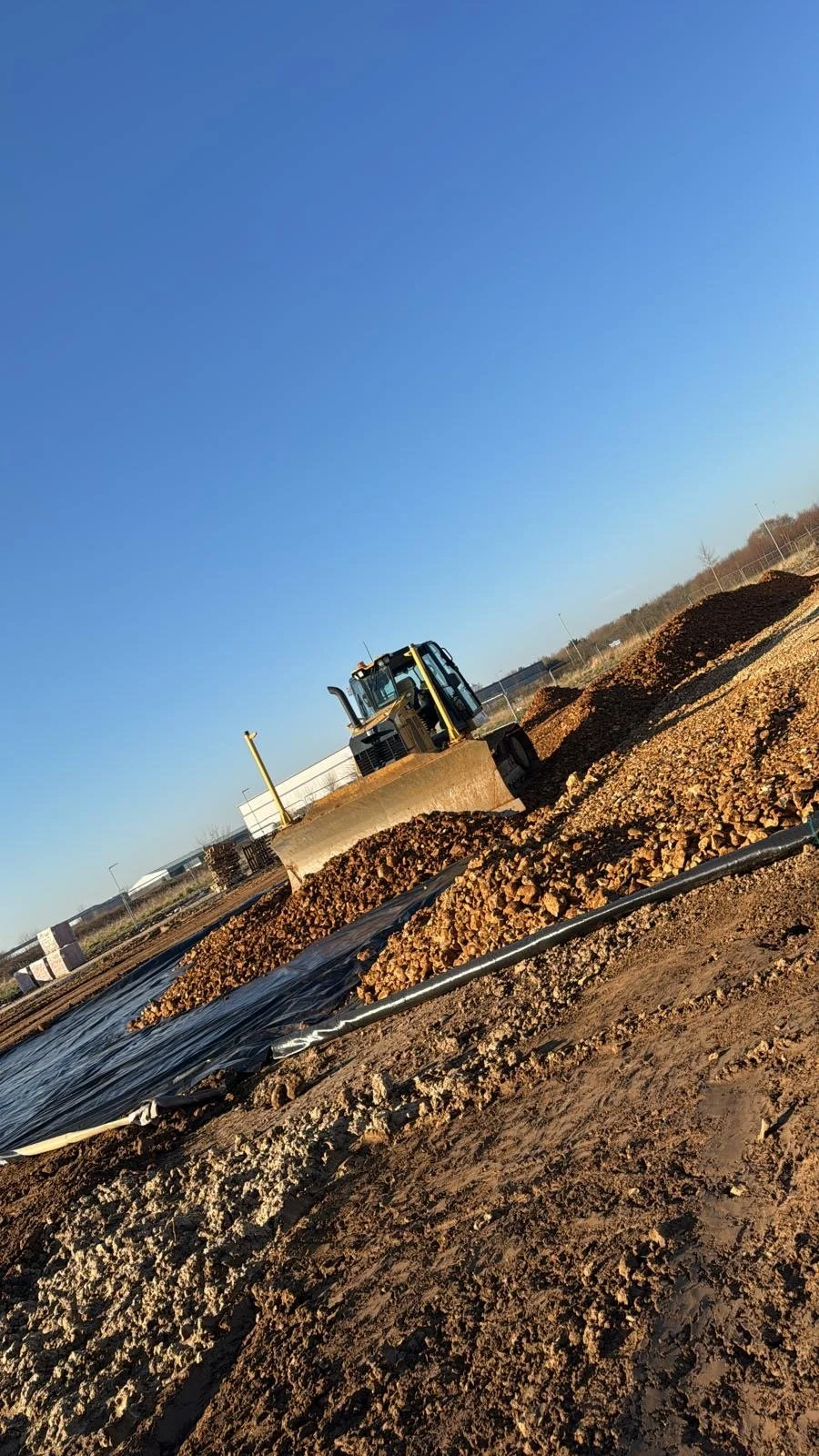 A construction site with a bulldozer moving earth and gravel, with clear blue sky and mounds of dirt in the background.