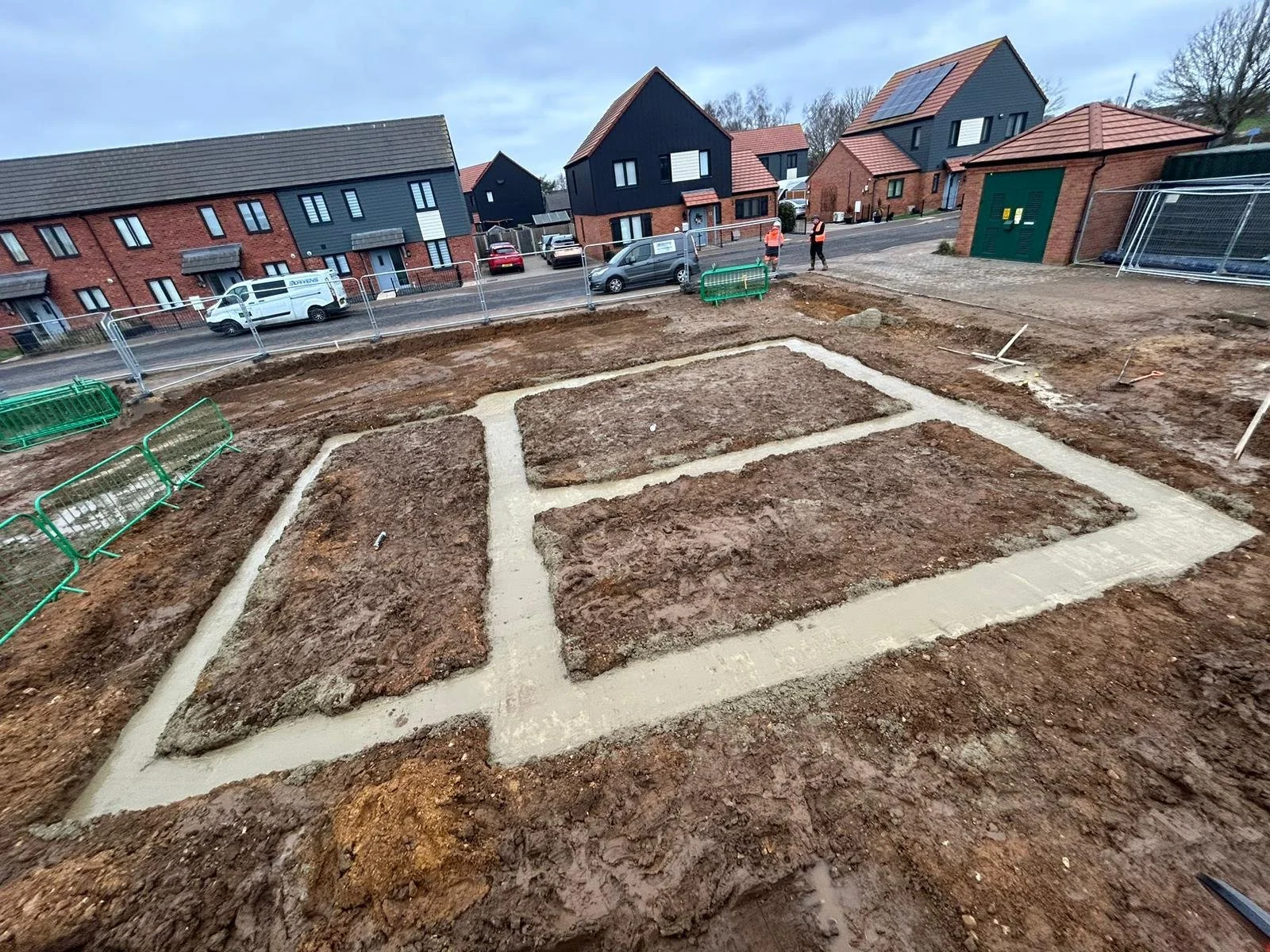 Construction site with cleared and excavated land, concrete footings or foundations outlined, residential houses in the background, workers in safety vests, and fencing around the area.