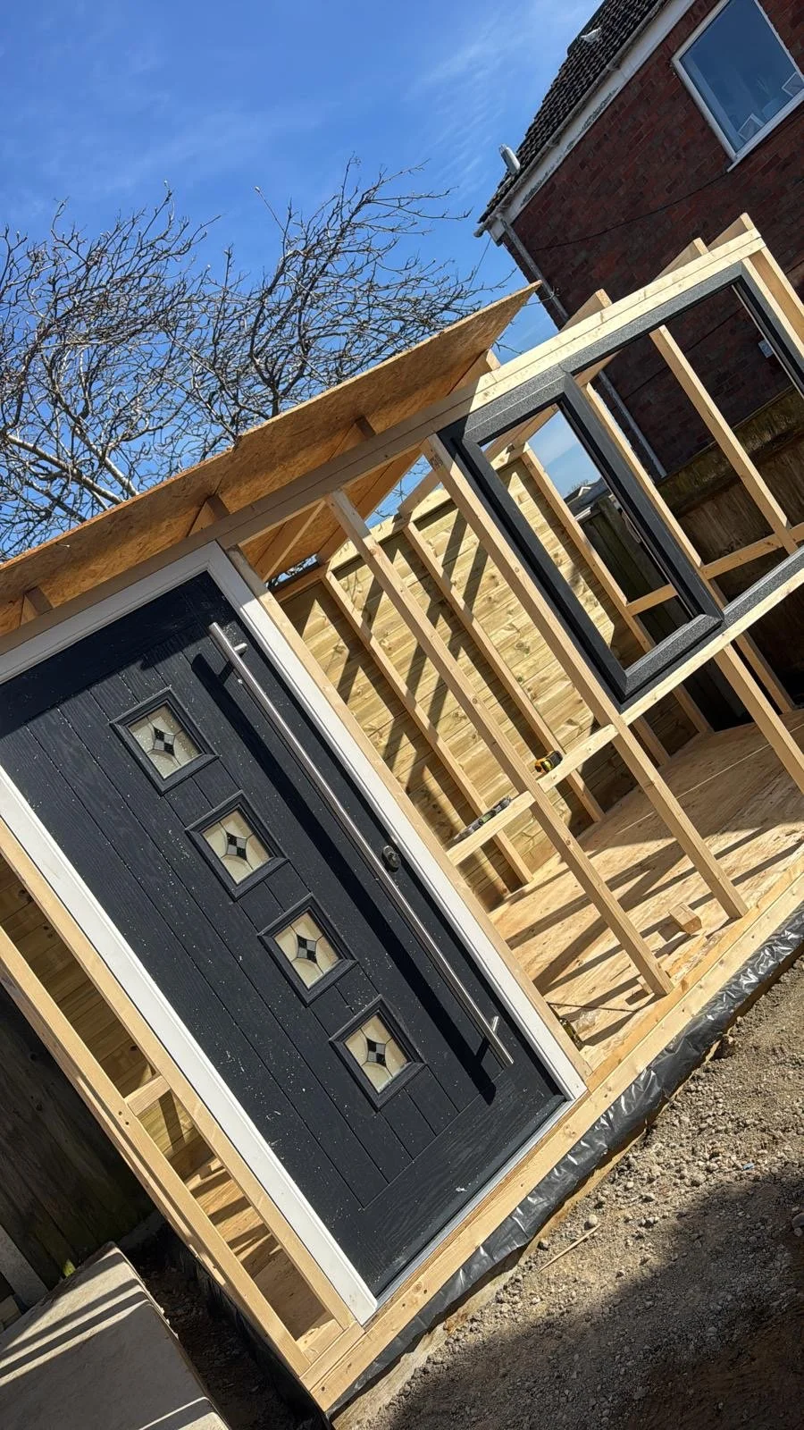 A building under construction with a black garage door featuring four small windows, wooden framing for a new extension, and a window already installed, with a clear blue sky and a leafless tree in the background.