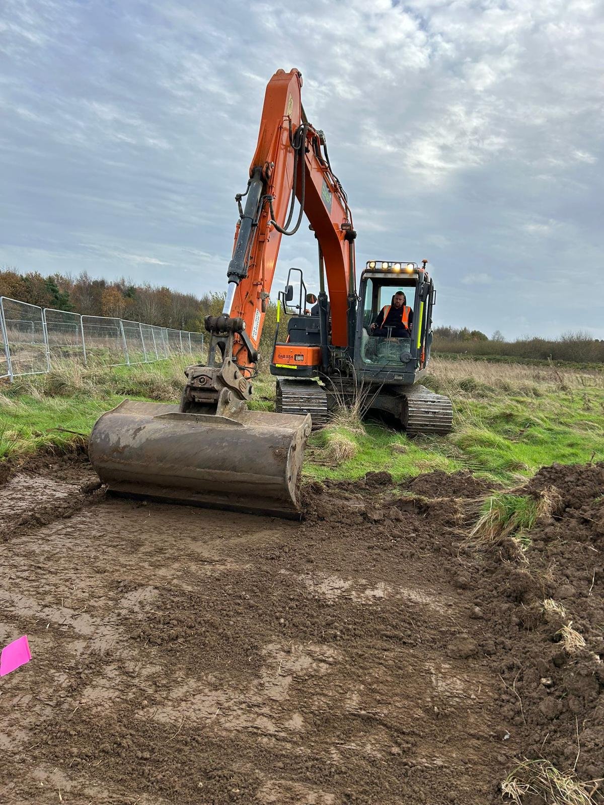 An orange excavator working on a construction site with a person operating it, moving dirt and earth, under a cloudy sky.