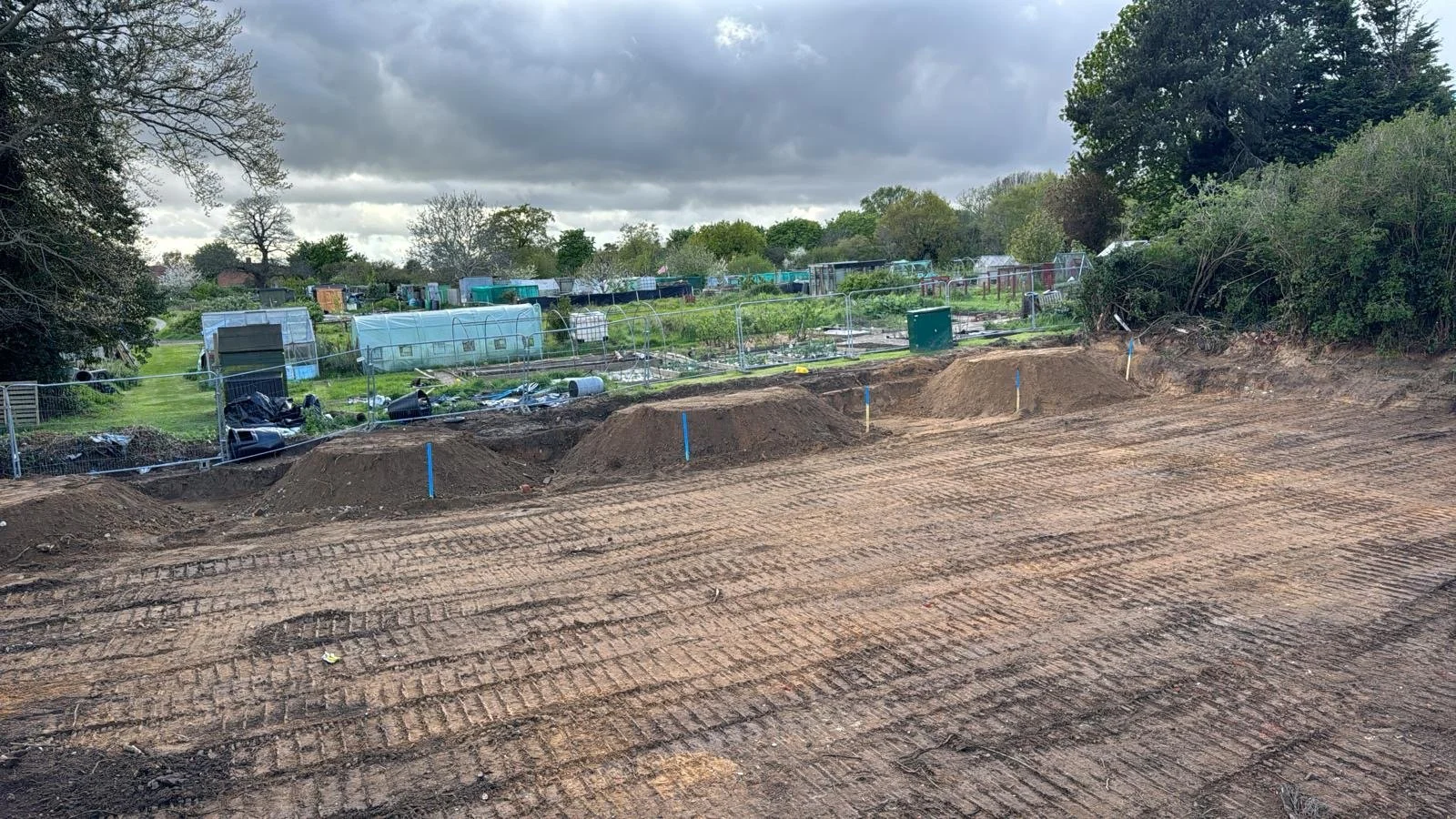 A construction site with dirt piles and construction marks in the foreground, gardens with greenhouses and trees in the background under a cloudy sky.