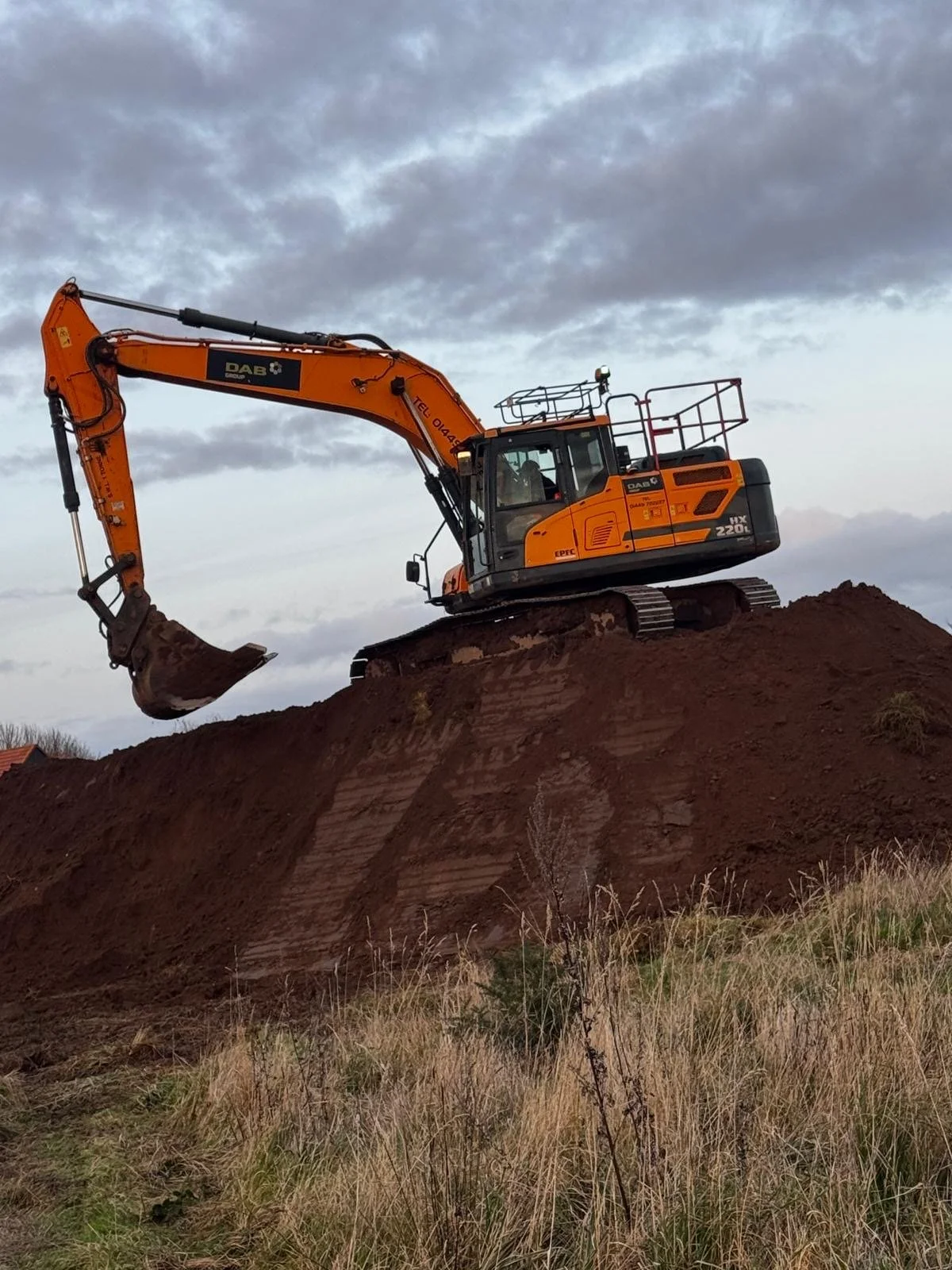 A large orange excavator on a mound of dirt with a cloudy sky in the background.
