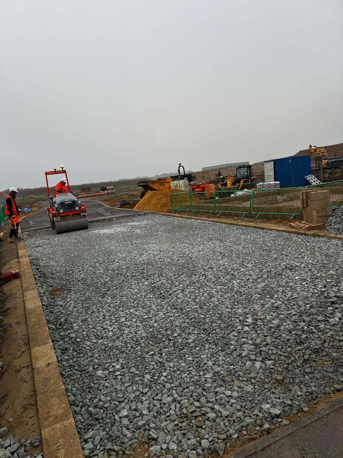 Construction workers paving a road with a roller compactor, surrounded by construction machinery and equipment, in a cloudy outdoor setting.