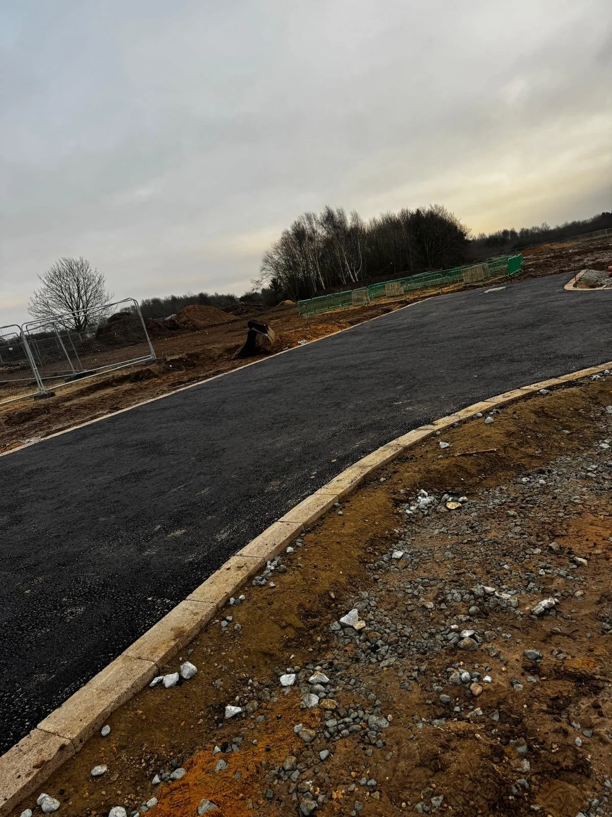 Newly paved asphalt road curving to the right on a construction site, with dirt and gravel on the sides, and construction barriers and trees in the background under a cloudy sky.