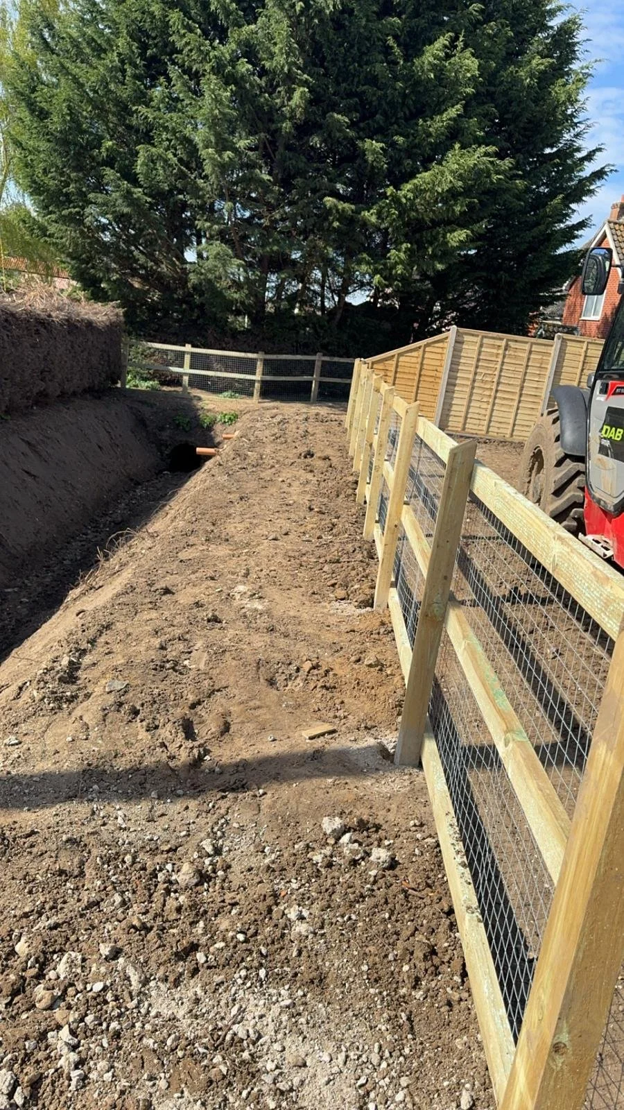 A newly dug trench with drainage beside a wooden and wire fence, with a large tree in the background and part of a red digger to the side on the right