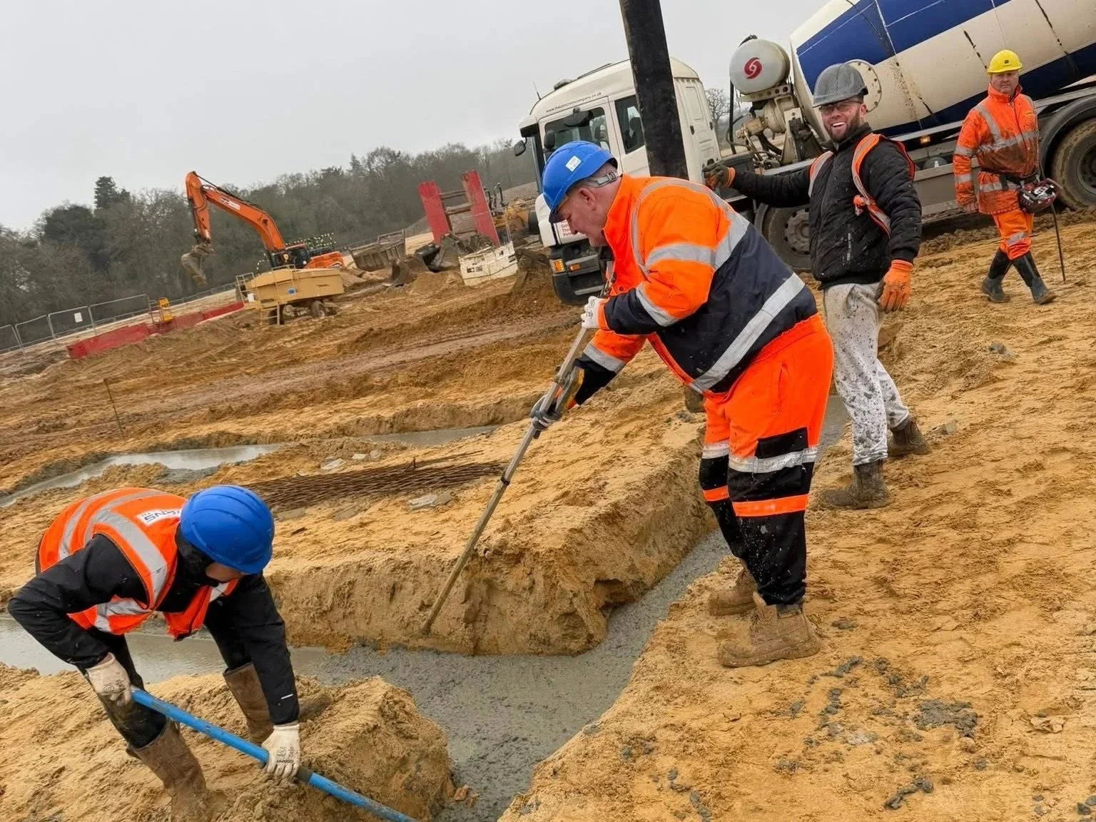 Construction workers in orange and blue safety gear are working on a sandy site, pouring concrete. Heavy machinery, including an excavator and cement mixer truck, are visible in the background.
