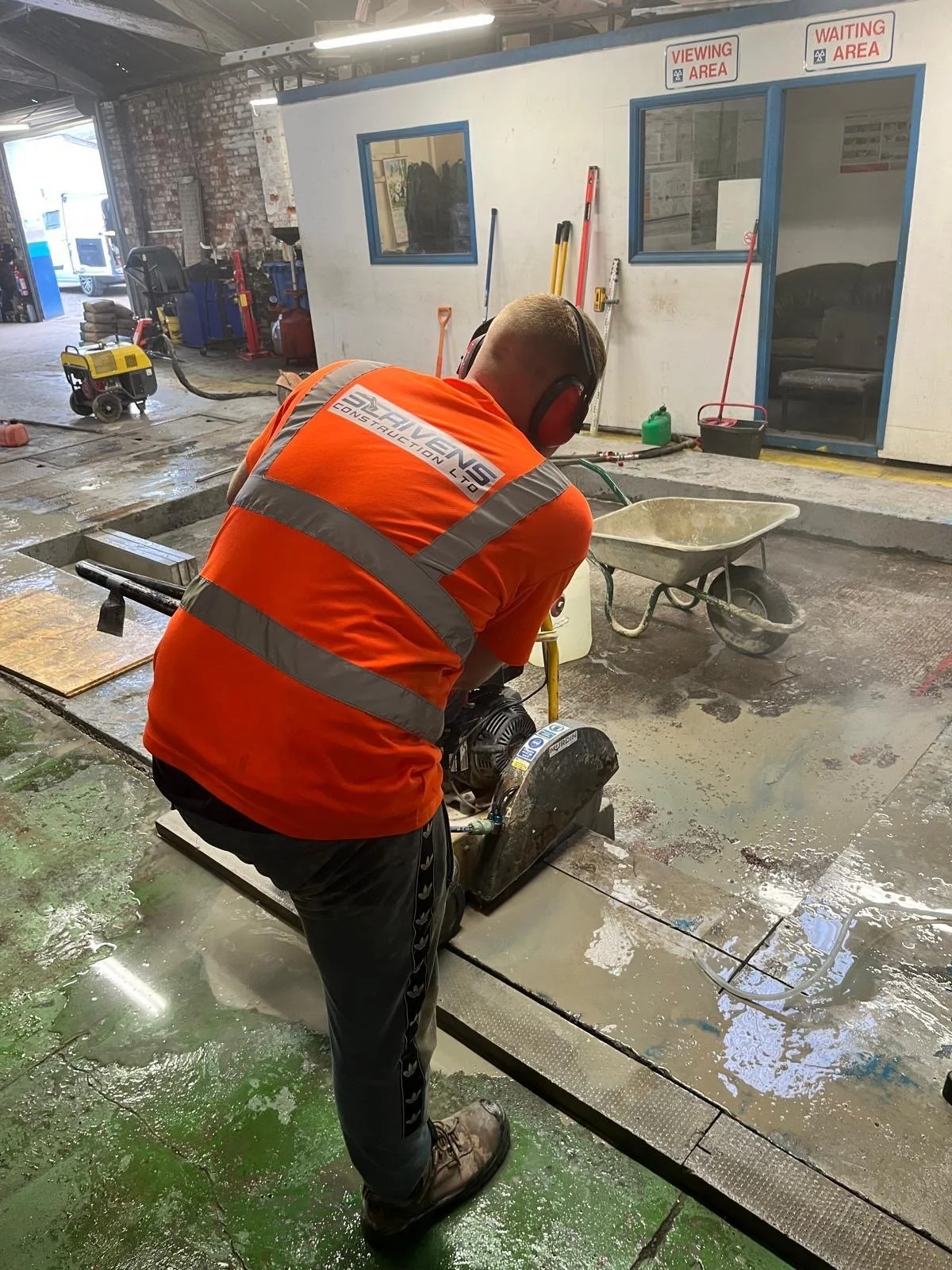Construction worker in orange safety shirt and ear protection cutting concrete with a floor saw in an indoor construction area.