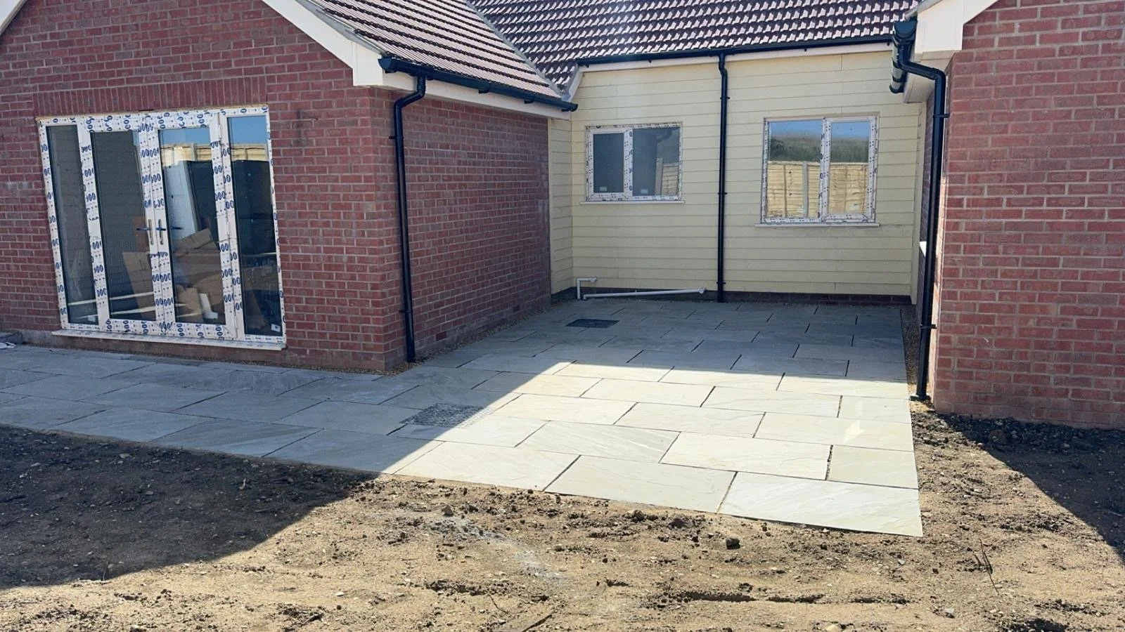 Newly laid stone patio in front of a house with red brick and cream siding, with black gutters and drainpipe, and unfinished soil surrounding the area.