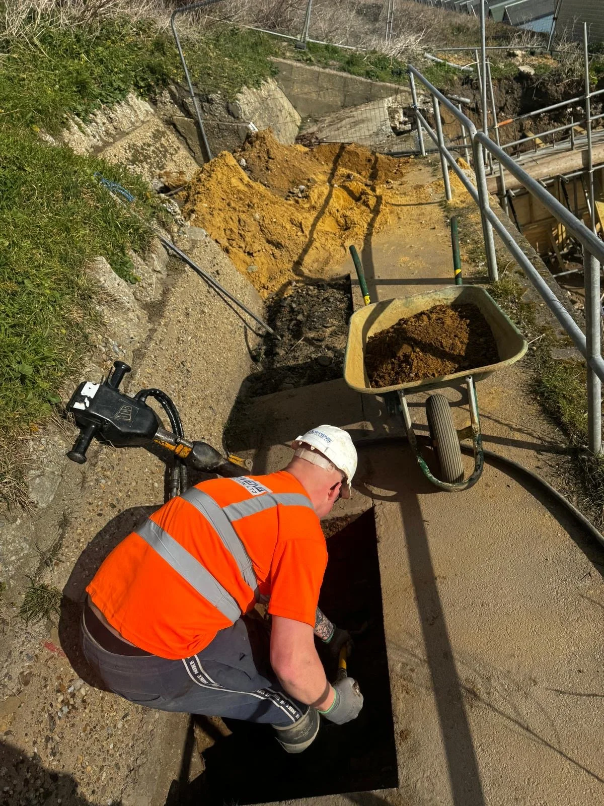 A construction worker in an orange safety vest and white hard hat is working at a dug-up section of promenade, with a wheelbarrow filled with dirt nearby and stairs with metal railing in the background.