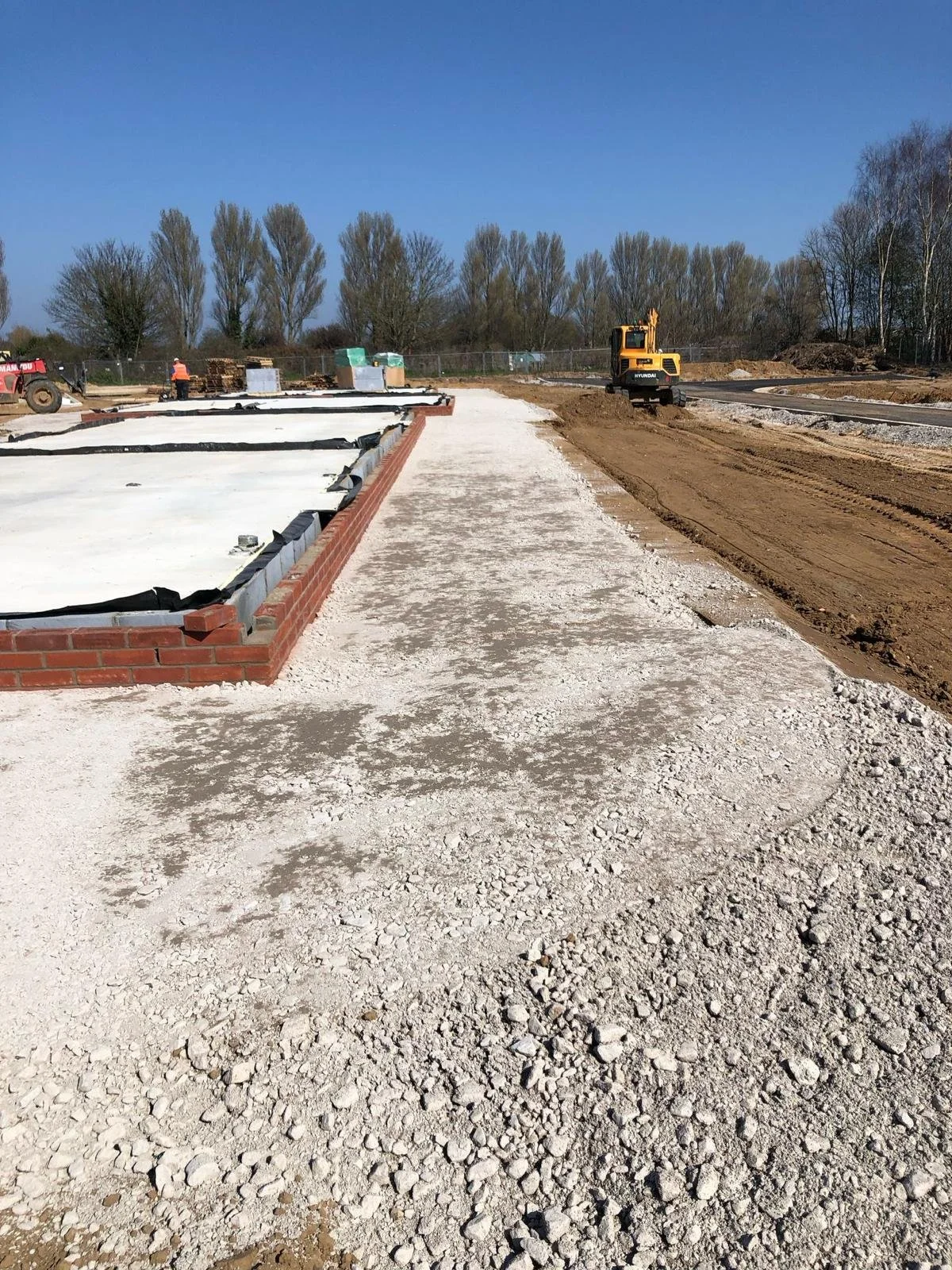 Construction site with a brick-bordered foundation, gravel pathway, and a small excavator, with workers and trees in the background under a clear blue sky.