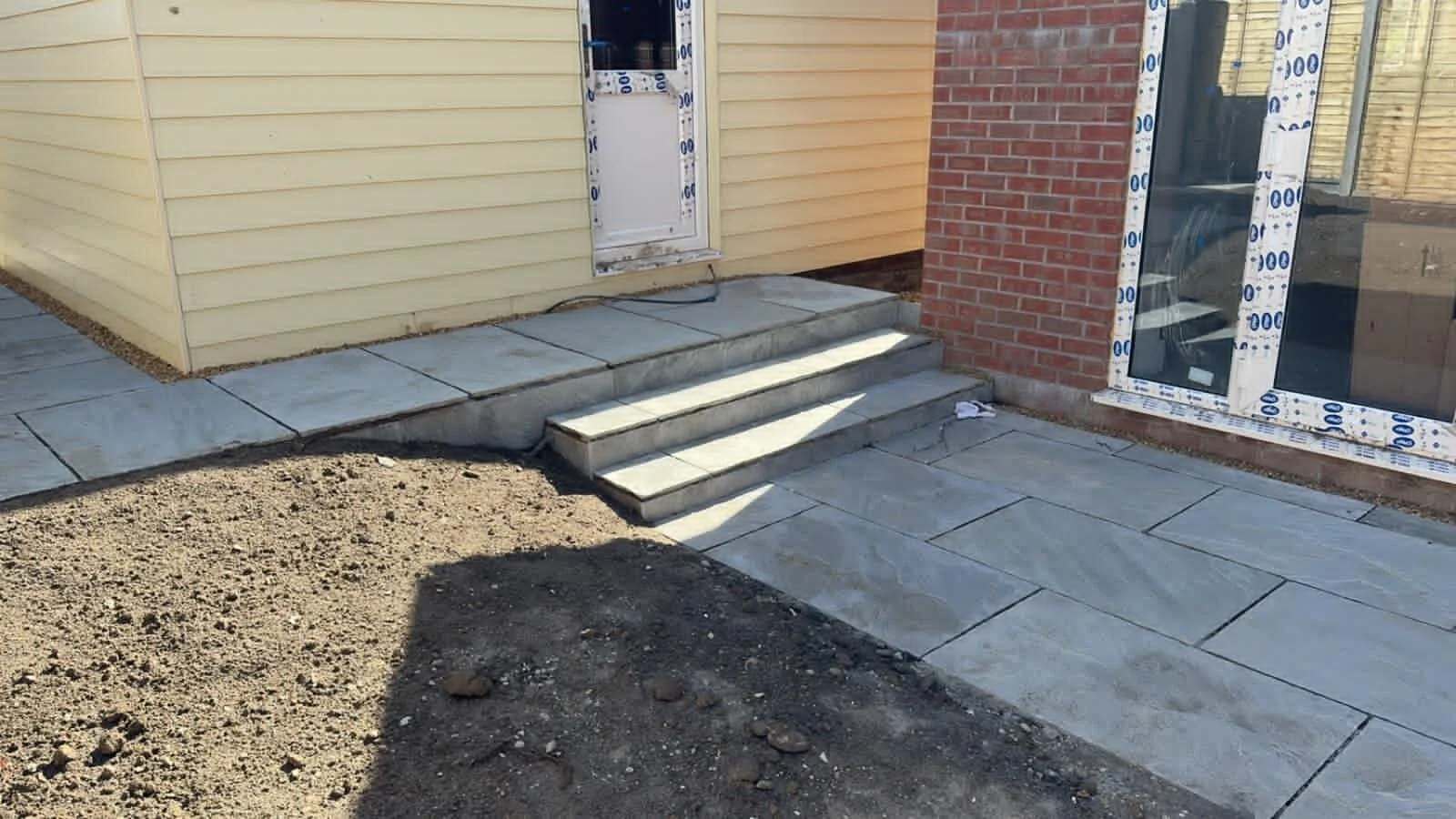 Newly installed concrete steps leading to a door on a house with beige siding, red brick wall, and sliding glass door, with a stone-paved patio area and dirt patch in the foreground.