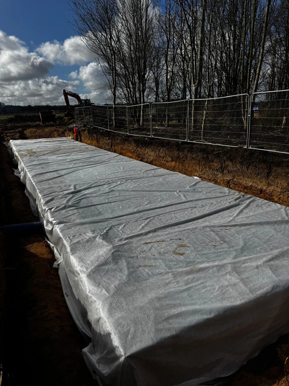 A construction site with a large trench covered with white geotextile fabric, surrounded by a metal fence, and an excavator in the background under a partly cloudy sky.