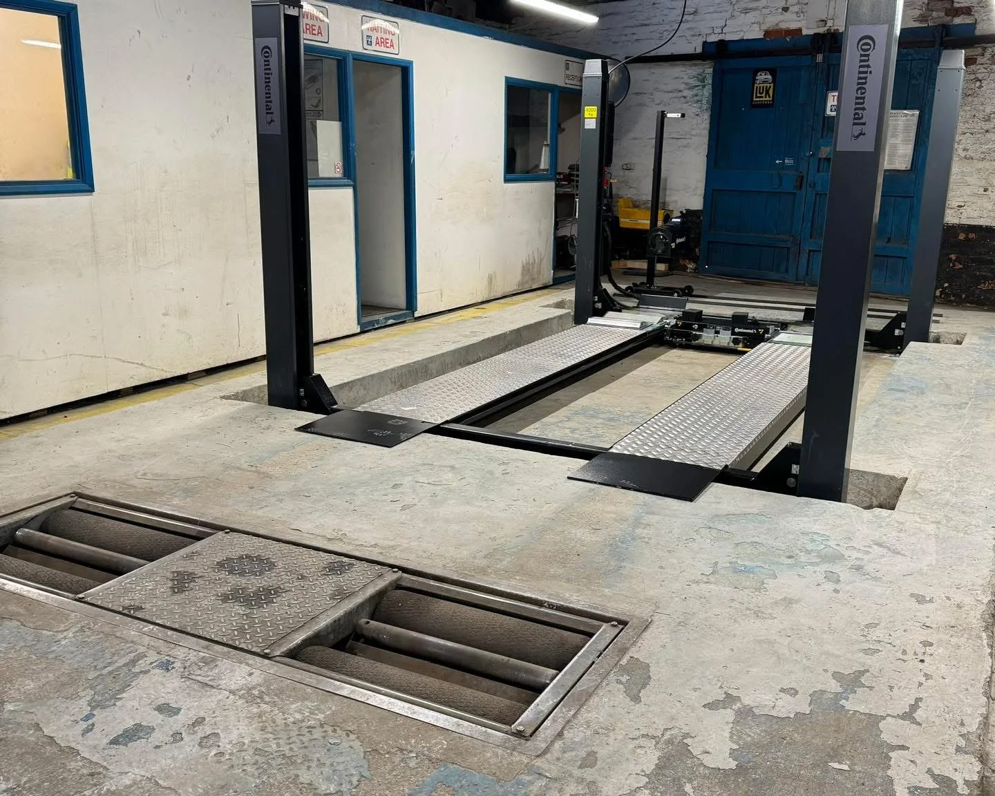 Automotive vehicle lift in a garage, with a drain on the concrete floor in the foreground and a blue storage container in the background.