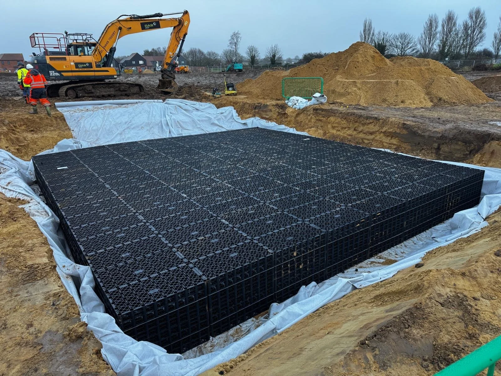 Construction site with a large black plastic soak-away structure installed in a dug-out area, workers and heavy machinery in the background, and piles of soil.
