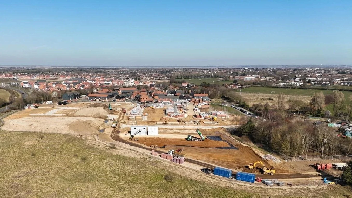 Construction site with machinery and foundation work in foreground, residential houses in background, trees on the right, and a clear blue sky.