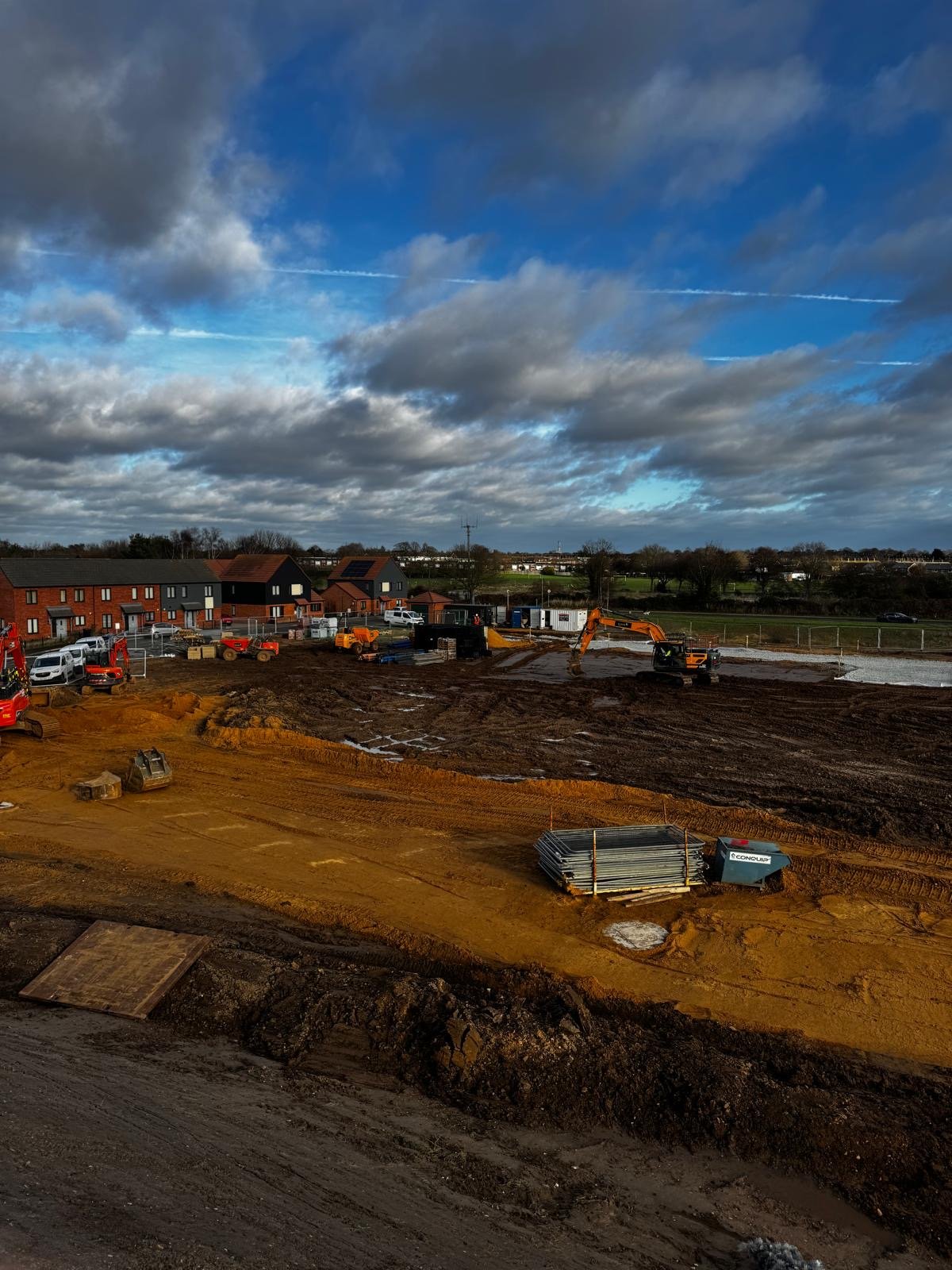 Construction site with excavators and dirt piles, with residential houses and a cloudy sky in the background.