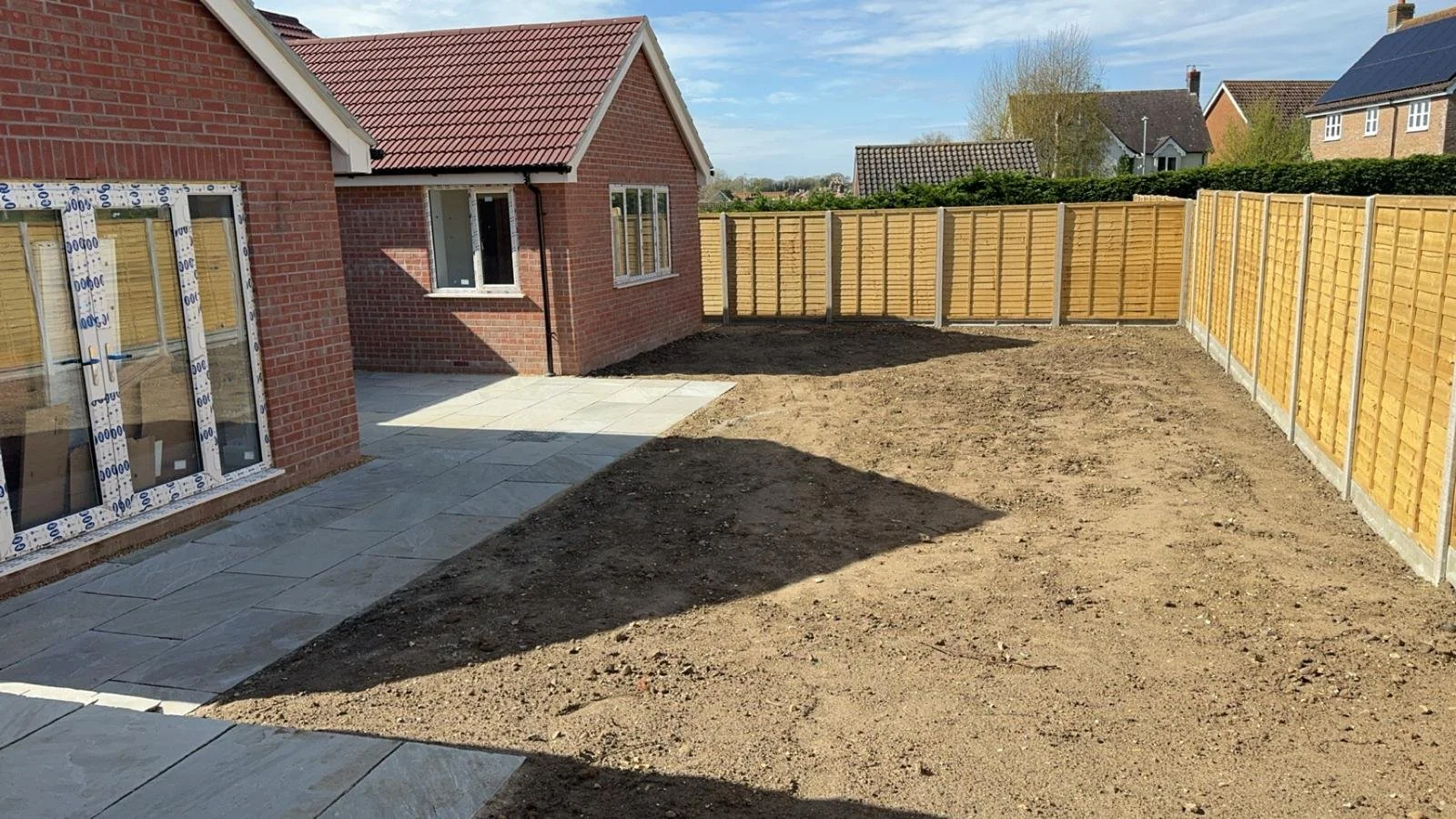 Backyard of a house with a tiled patio area, partially cleared land, and wooden fencing, under a clear blue sky.