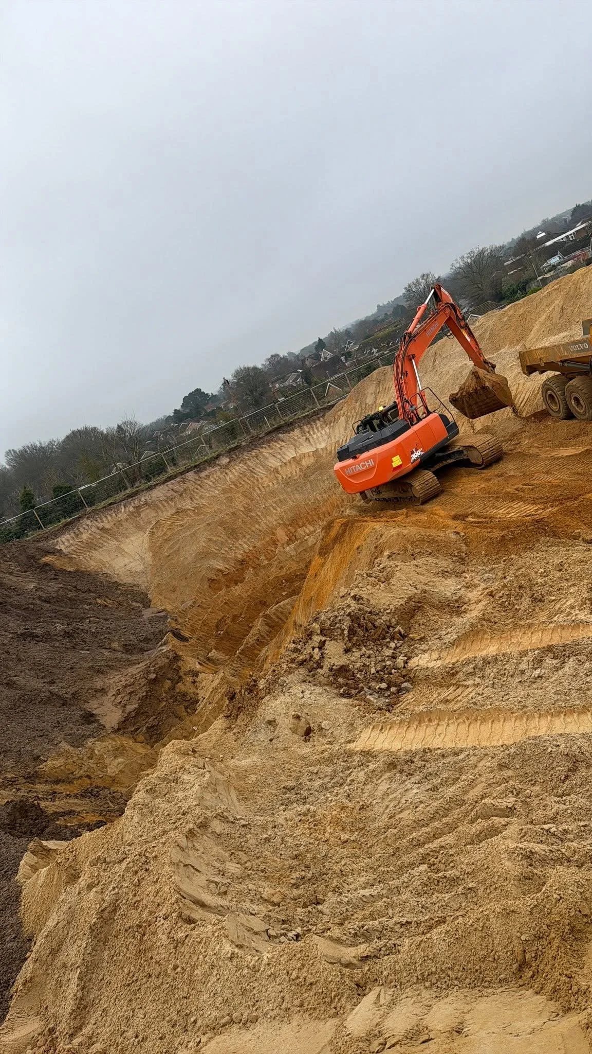 An orange excavator working on a sandy construction site, moving dirt with a large bucket, with a hill and buildings in the background.