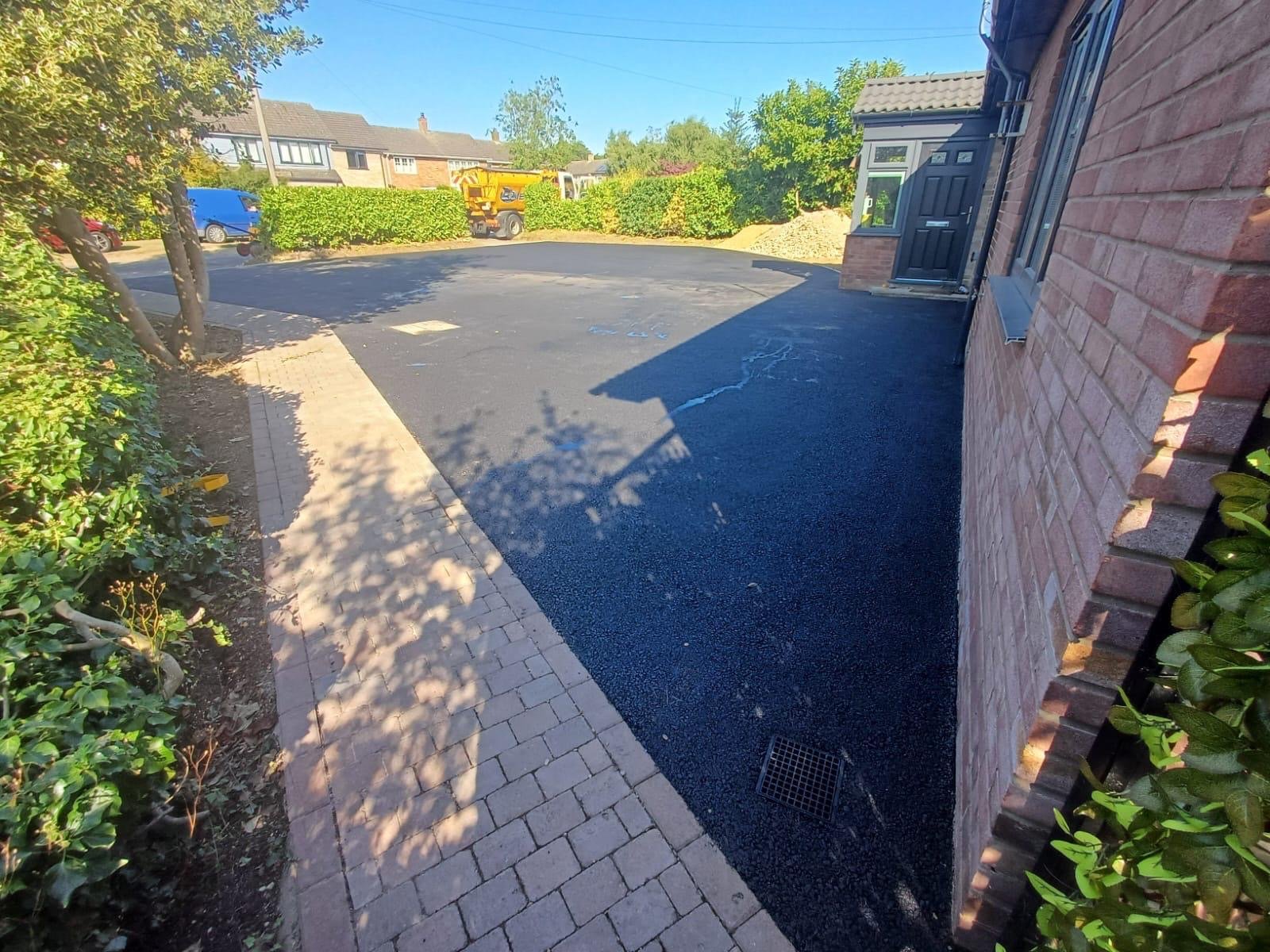 A newly paved asphalt driveway next to a brick house, with a small brick walkway and garden bed on the left, and a drain grate near the house on the right.