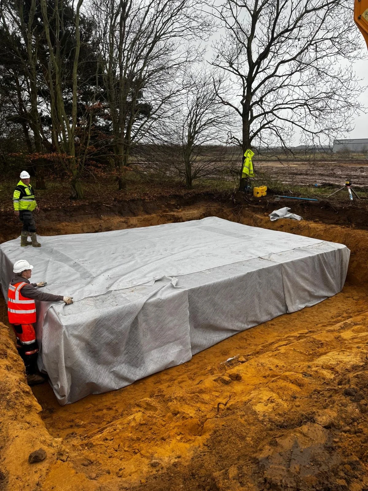 Construction workers installing a large underground utility vault (soak away) covered with a tarp at a construction site surrounded by soil and trees.