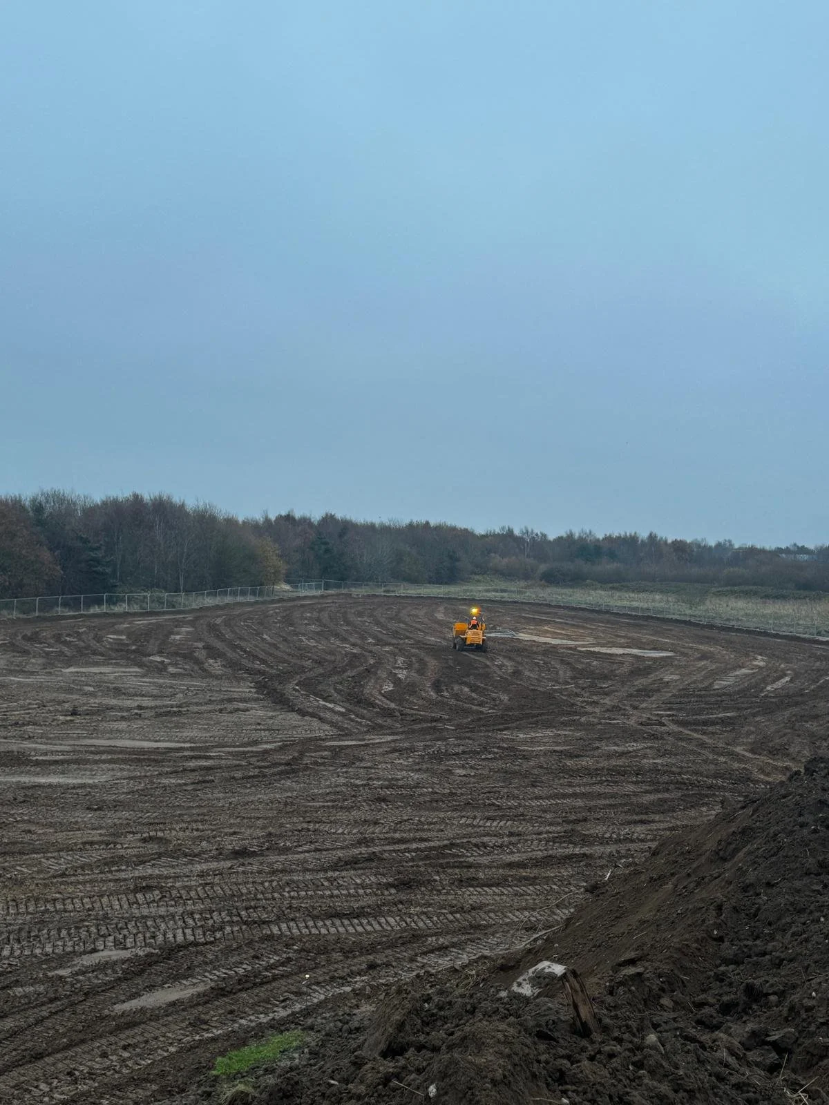 A wide view of a construction site with uneven dirt ground and tire tracks, and a small yellow construction vehicle operating in the distance under a cloudy sky.