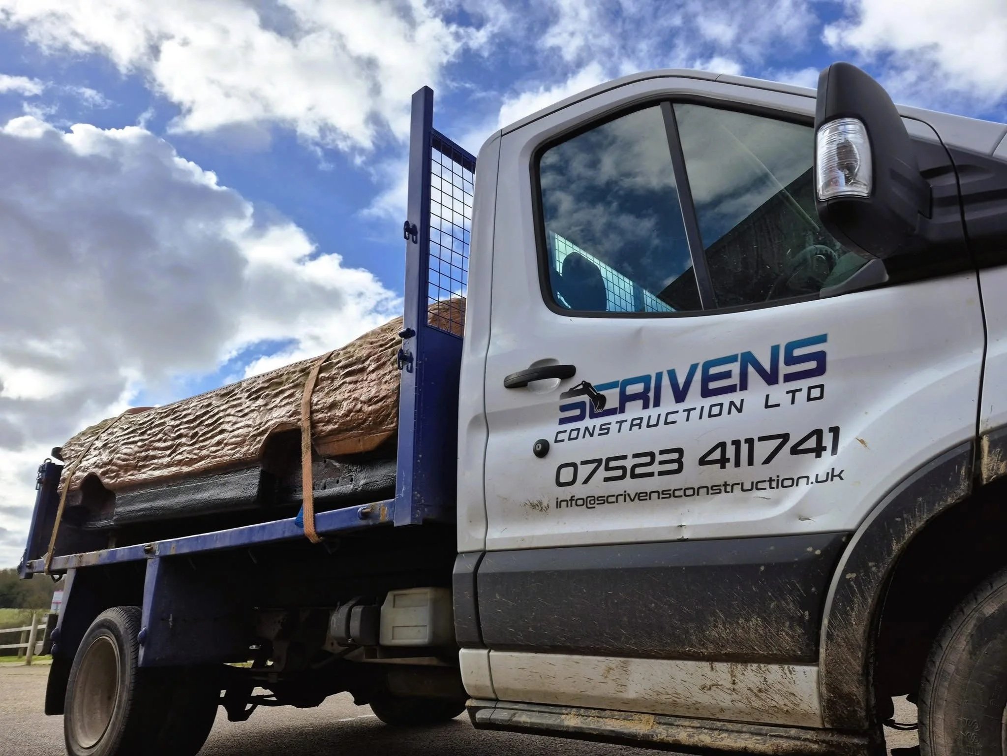 A flatbed truck carrying a large log with a wood grain visible, parked outdoors under a partly cloudy sky, with company branding for "Scrivens Construction Ltd," a phone number, and an email address on the side of the truck.