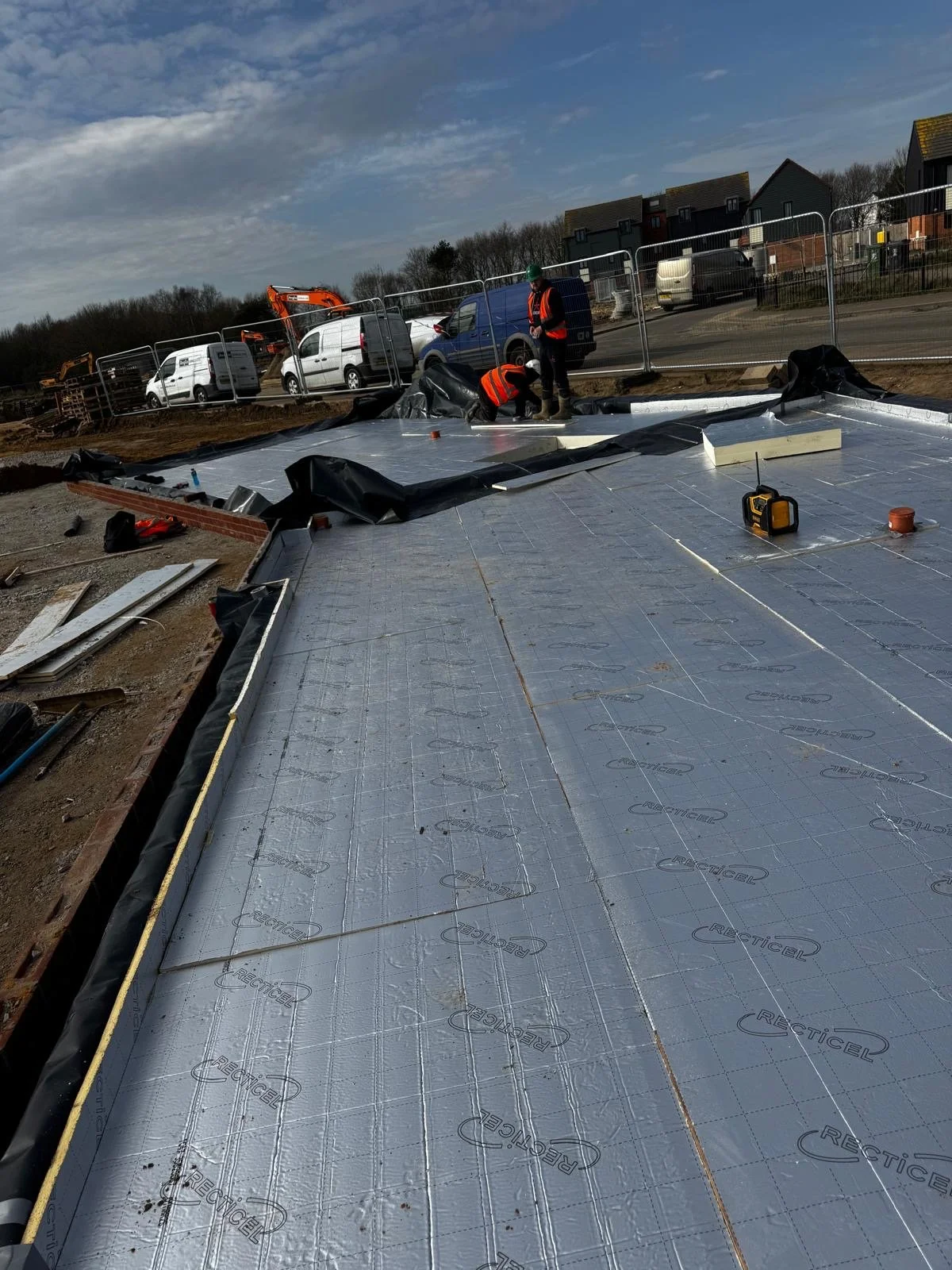 Construction workers installing waterproofing membrane on a building foundation in an outdoor construction site, with vehicles and residential buildings in the background.