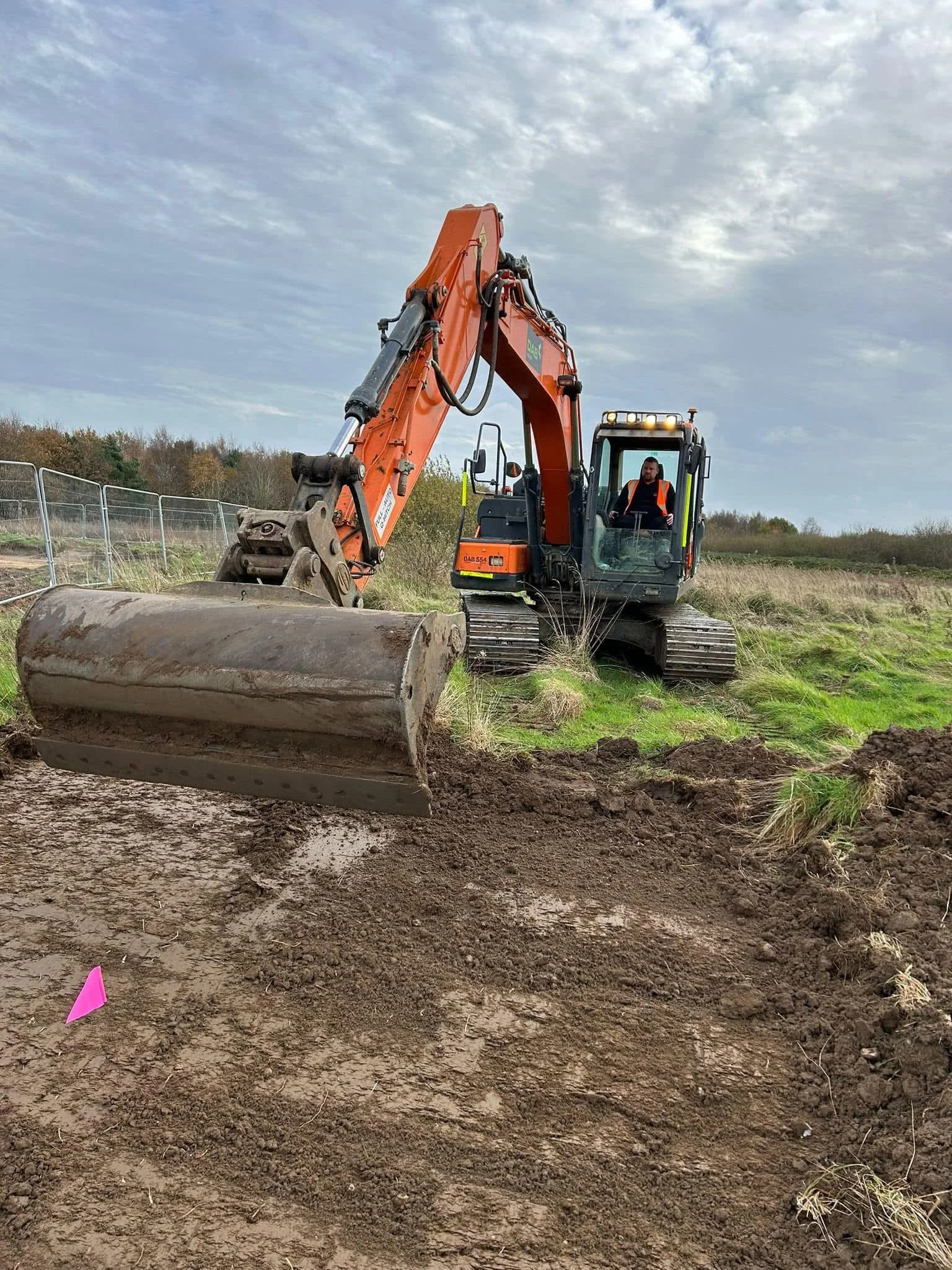 A person operating an orange track excavator on a grassy construction site, digging into the dirt with a large blade. The background shows a cloudy sky and a fenced area.