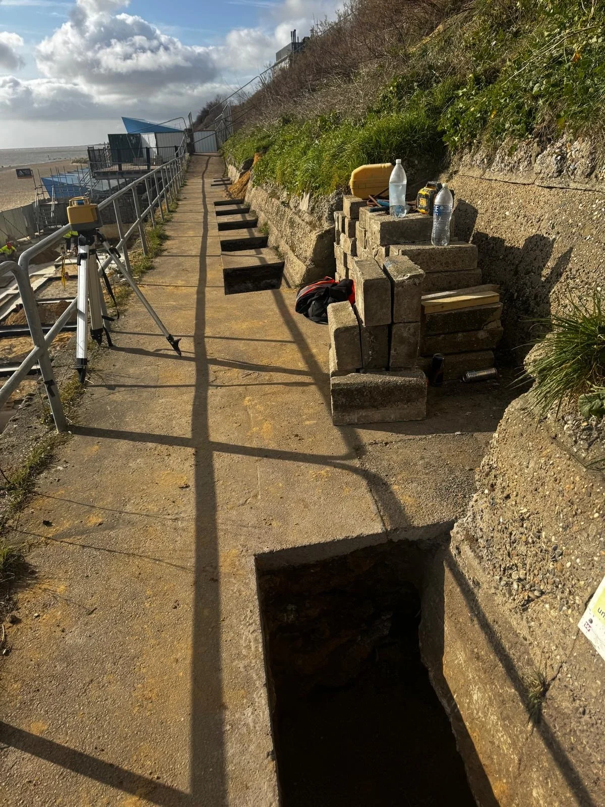 Pathway along a beach with construction or maintenance supplies needed for underpinning, including a tripod and stacked bricks, with a stairway leading up to a grassy area and a partly cloudy sky.