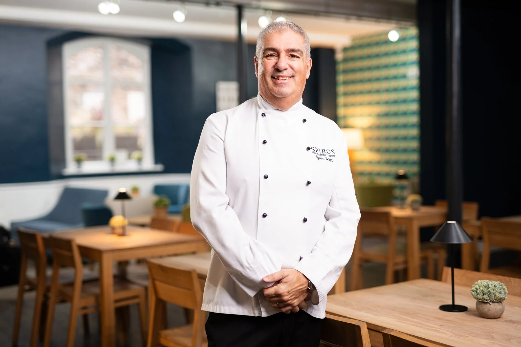 A smiling male chef wearing a white chef jacket standing in a modern restaurant with wooden tables and chairs, decorative lamps, and a window with natural light in the background.