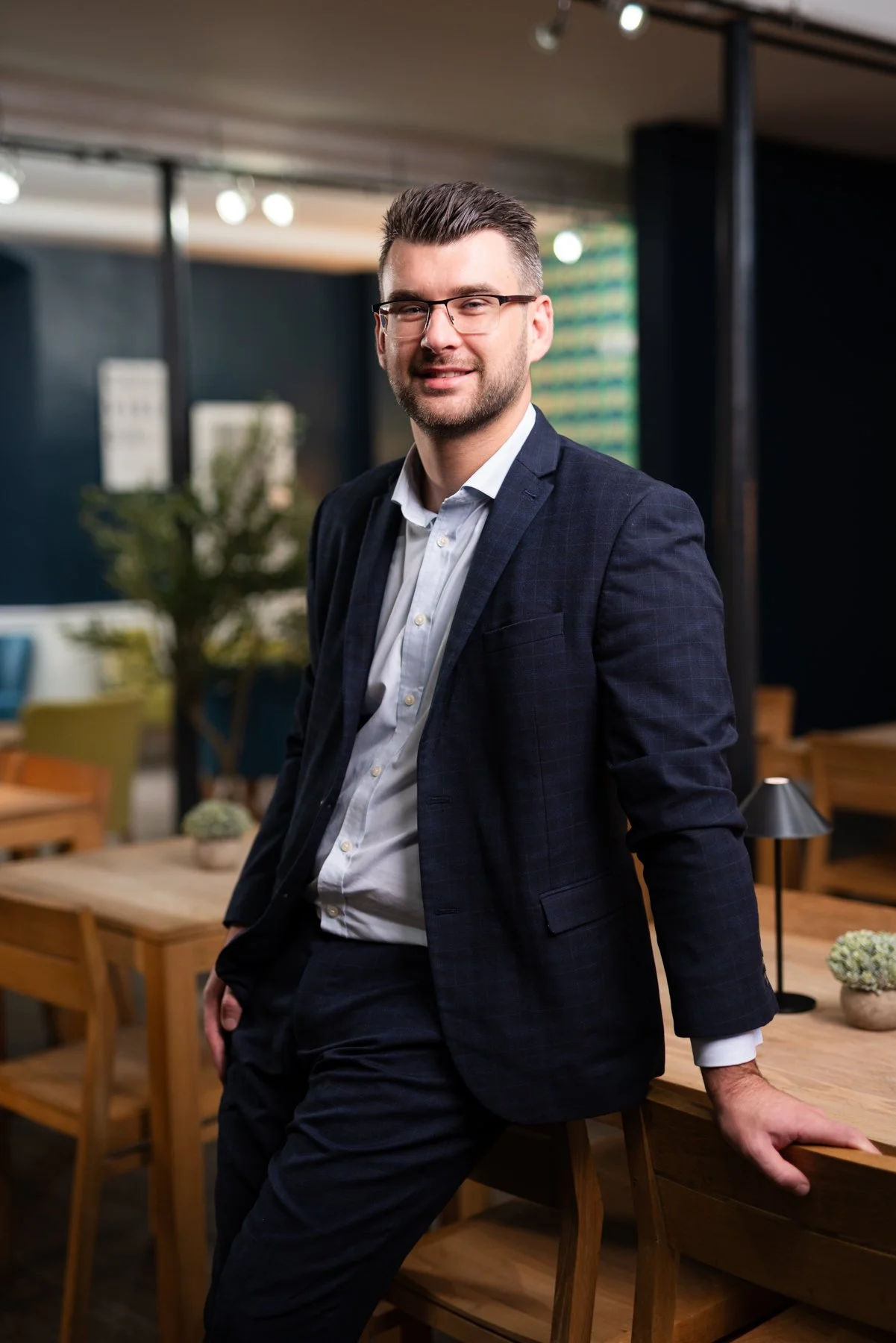 A man in a suit standing in an office or lounge area, leaning on a wooden table with a modern design, with blurred background including chairs, plants, and lighting fixtures.