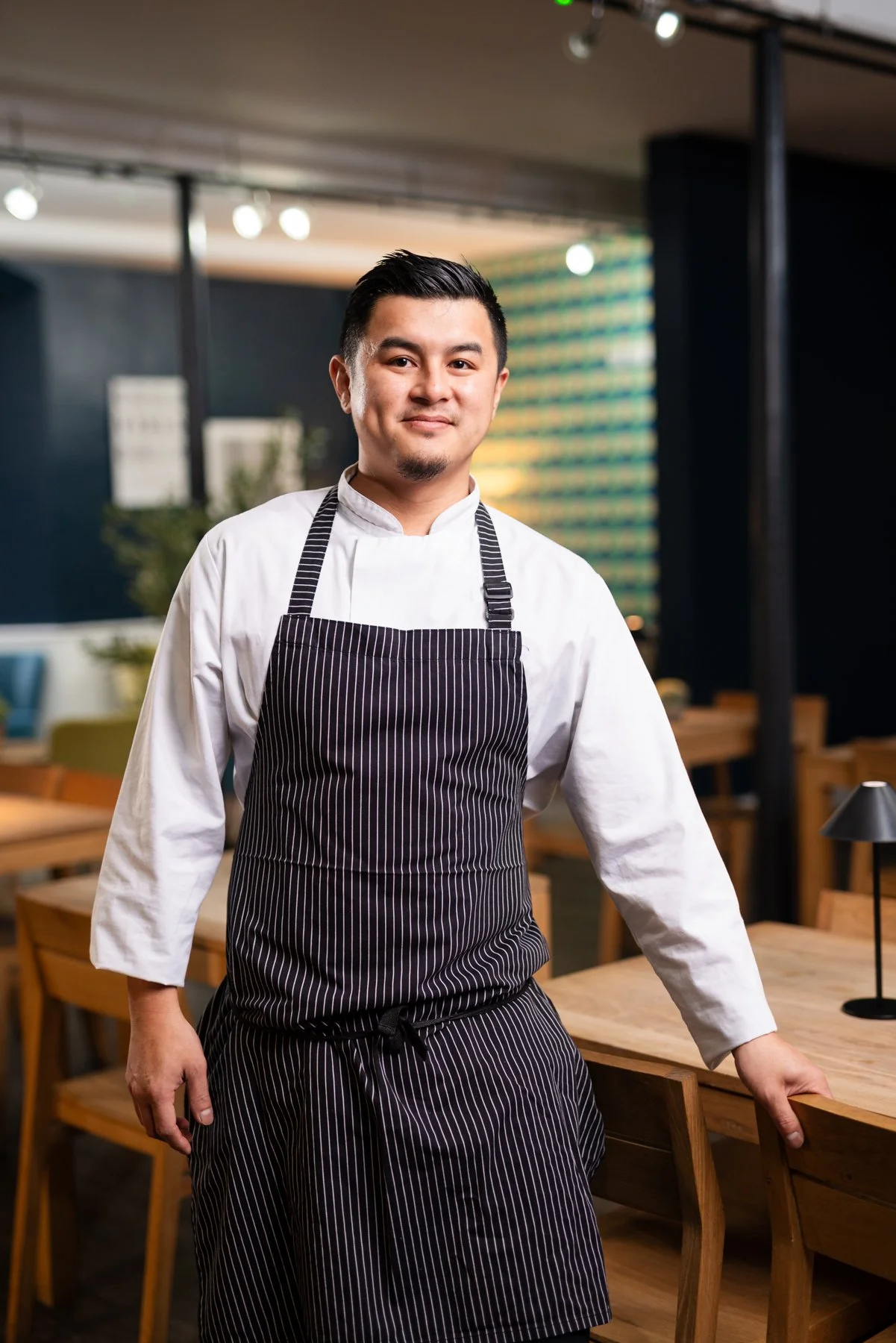 A male chef standing in a restaurant, wearing a white chef's coat and a black and white striped apron, smiling with a relaxed posture.