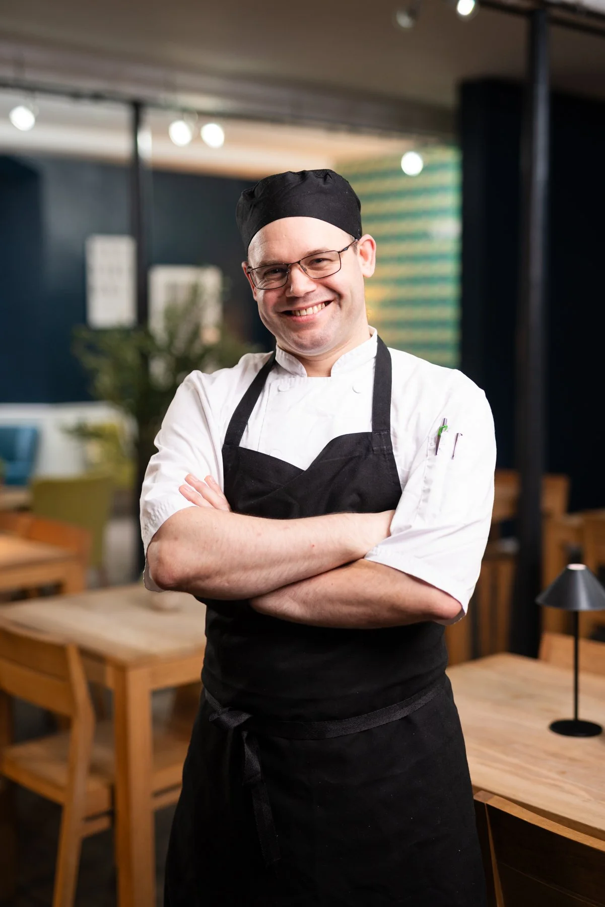 A smiling male chef wearing glasses, a black chef hat, a white chef's jacket, and a black apron, standing with arms crossed in a restaurant or cafe setting.