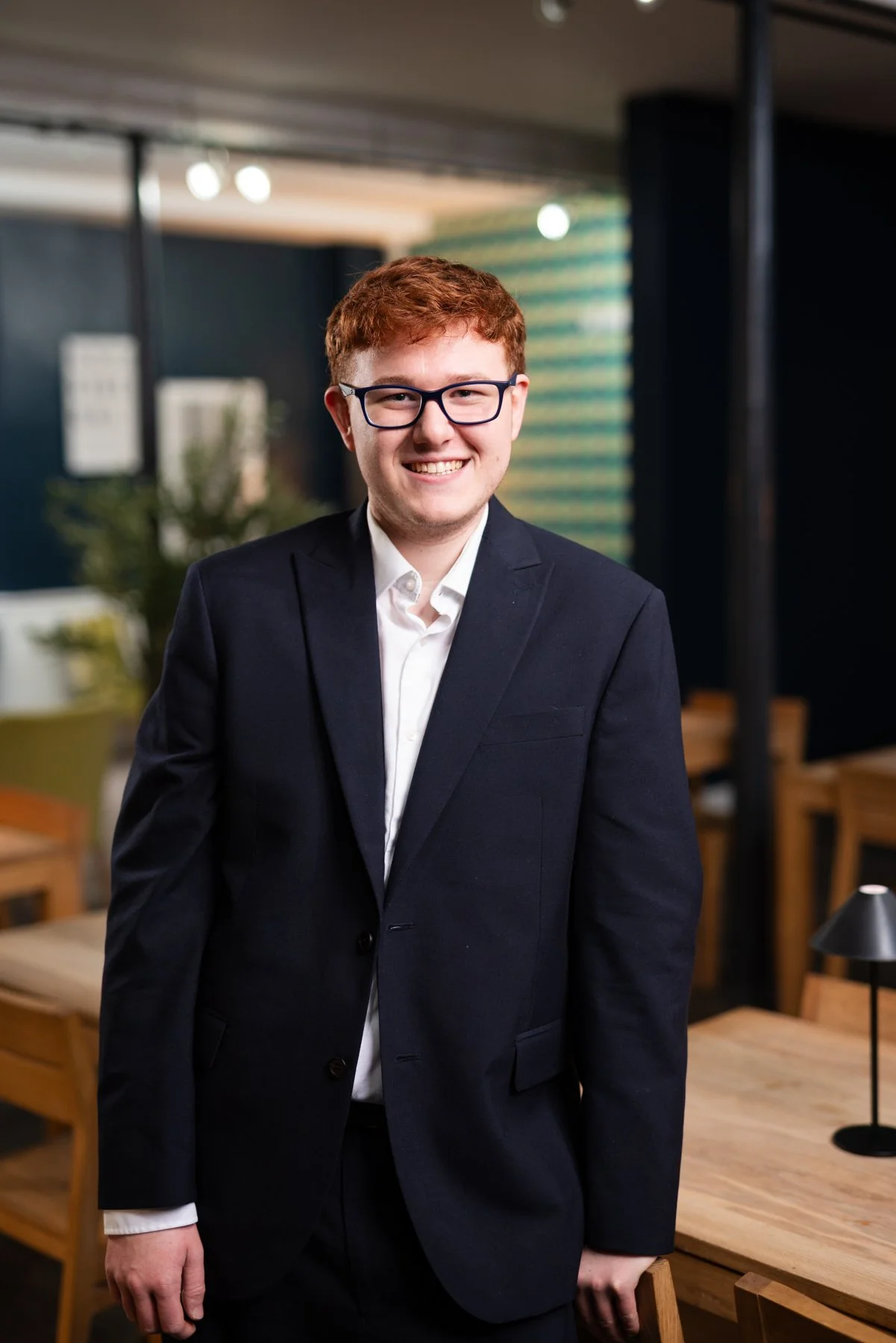 A young man with red hair, glasses, and a suit, smiling in an indoor setting with tables and chairs in the background.