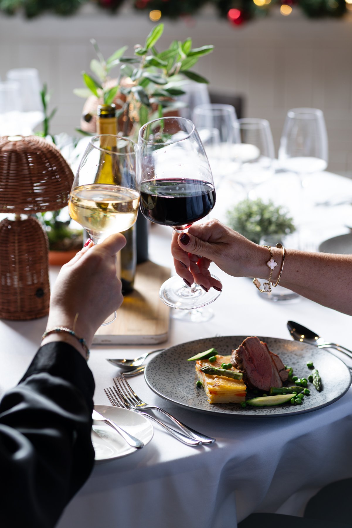 People clinking glasses of white and red wine over a dinner table with a plate of food, including meat, asparagus, and peas, decorated with a tablecloth and surrounded by glasses and the festive background.