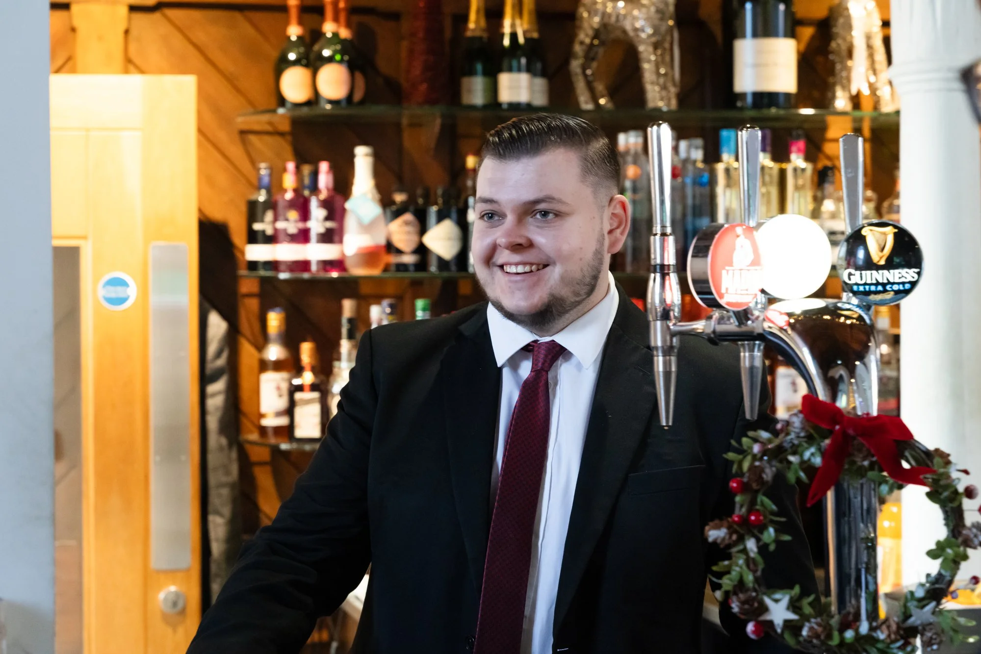 A young man with short, slicked-back hair, wearing a black suit, white shirt, and burgundy tie, smiling while working behind a bar with various bottles of alcohol and beer taps, in a warmly lit bar or pub setting.