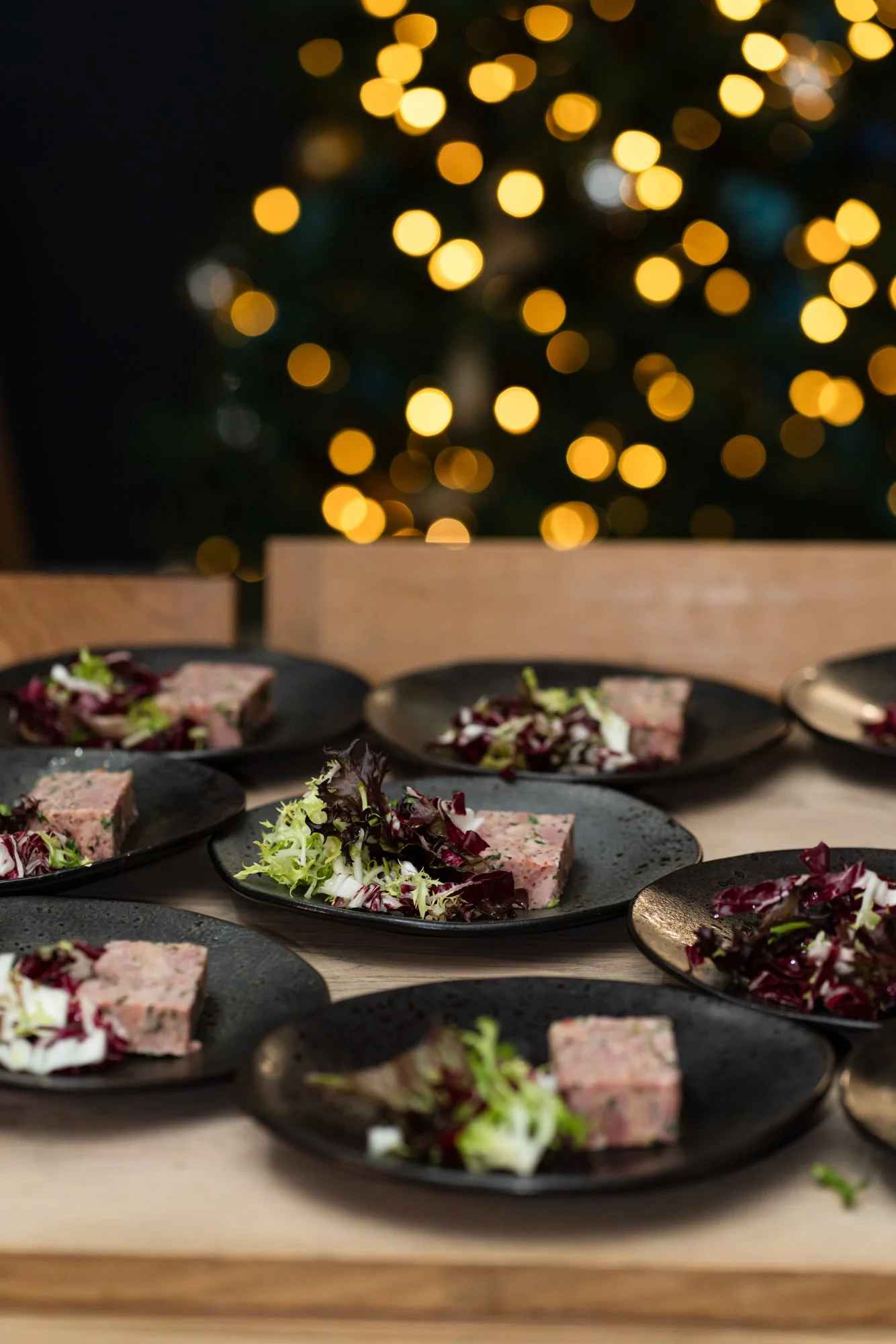 Plates of pâté garnished with mixed greens on a light wooden table, with blurred holiday lights in the background.
