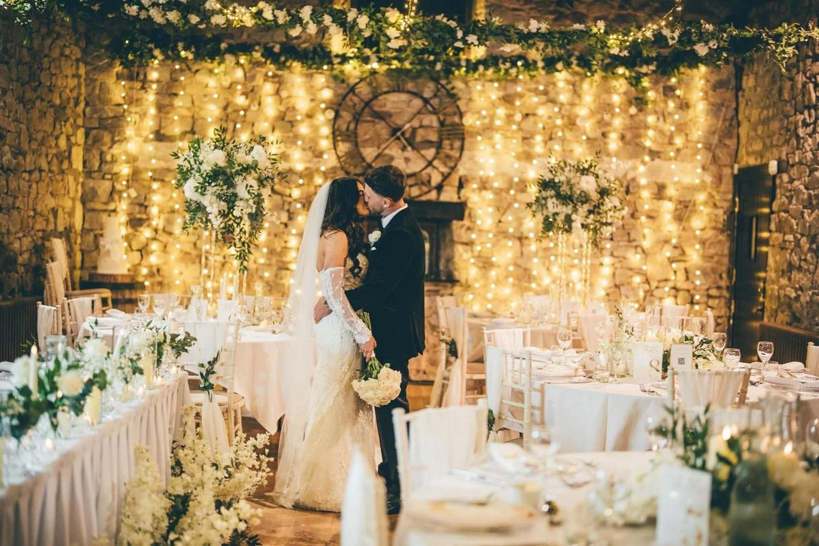 A bride and groom share a kiss at their wedding reception, decorated with string lights, large floral arrangements, and elegant table settings in a rustic stone venue.