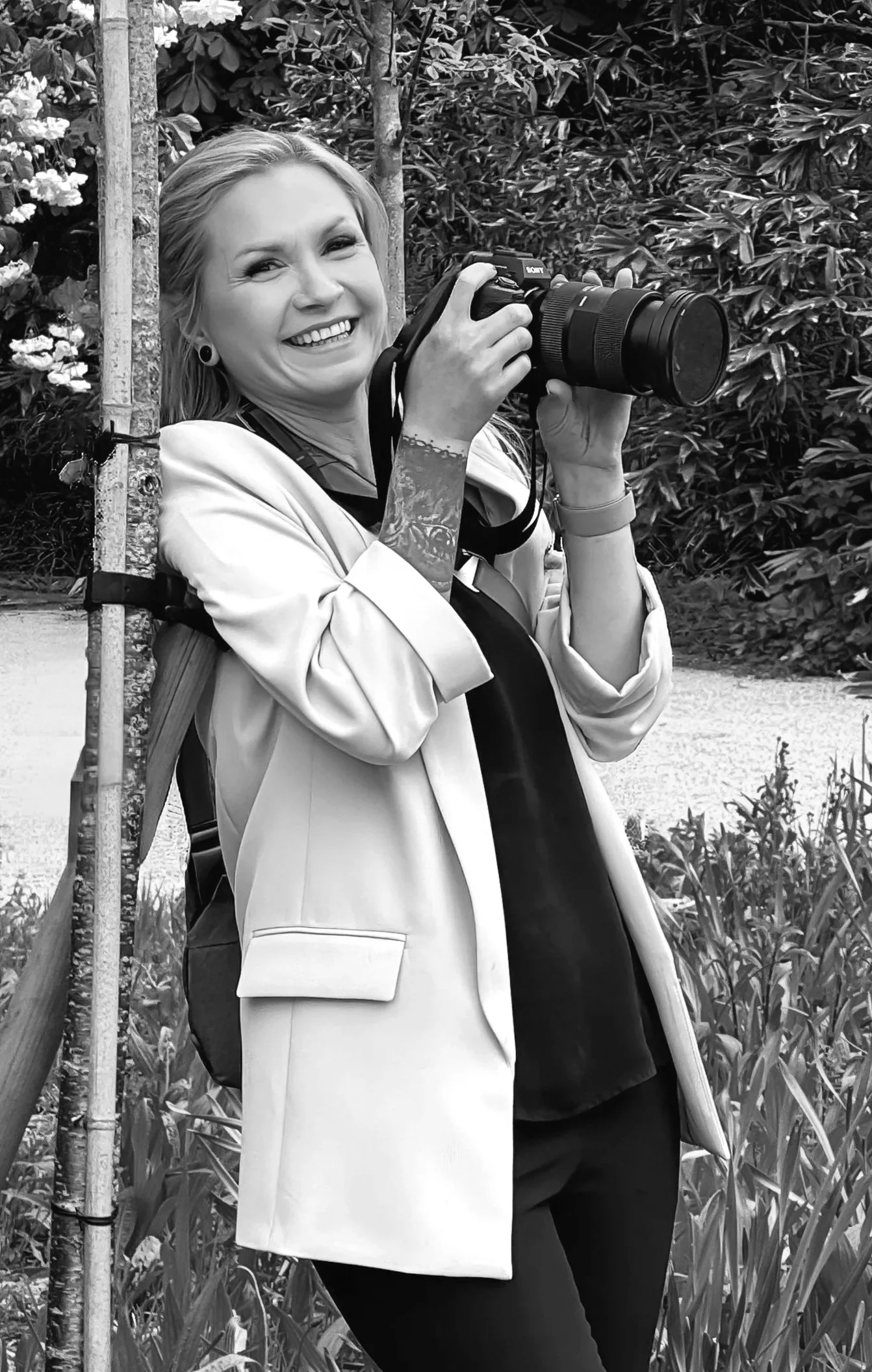 Photographer smiling and holding a camera, standing outdoors among plants with a tree behind her.