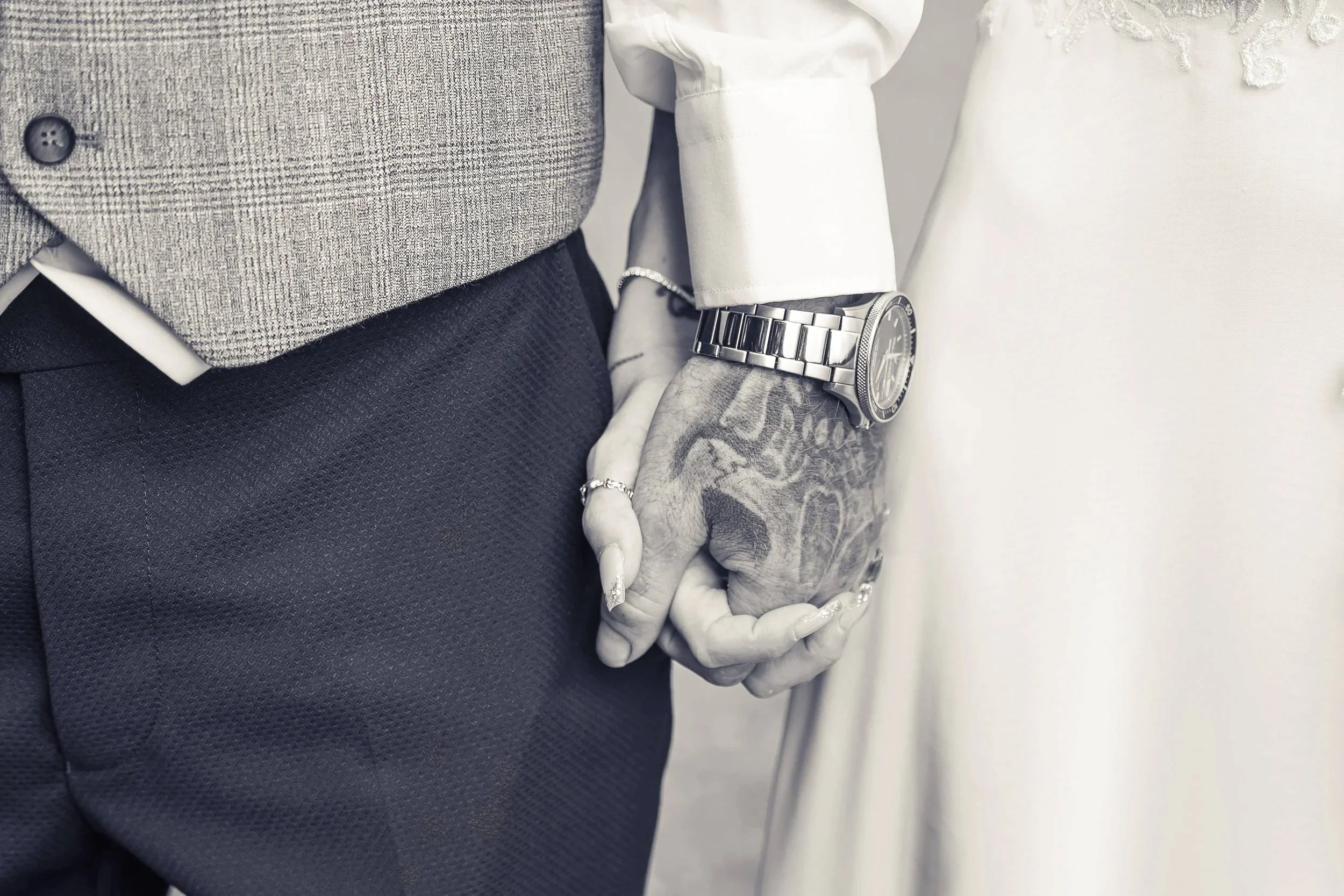 A close-up of a couple holding hands at their wedding, with the bride wearing a white dress and the groom in a gray suit. The groom has a wristwatch and the bride has decorated nails and jewelry.