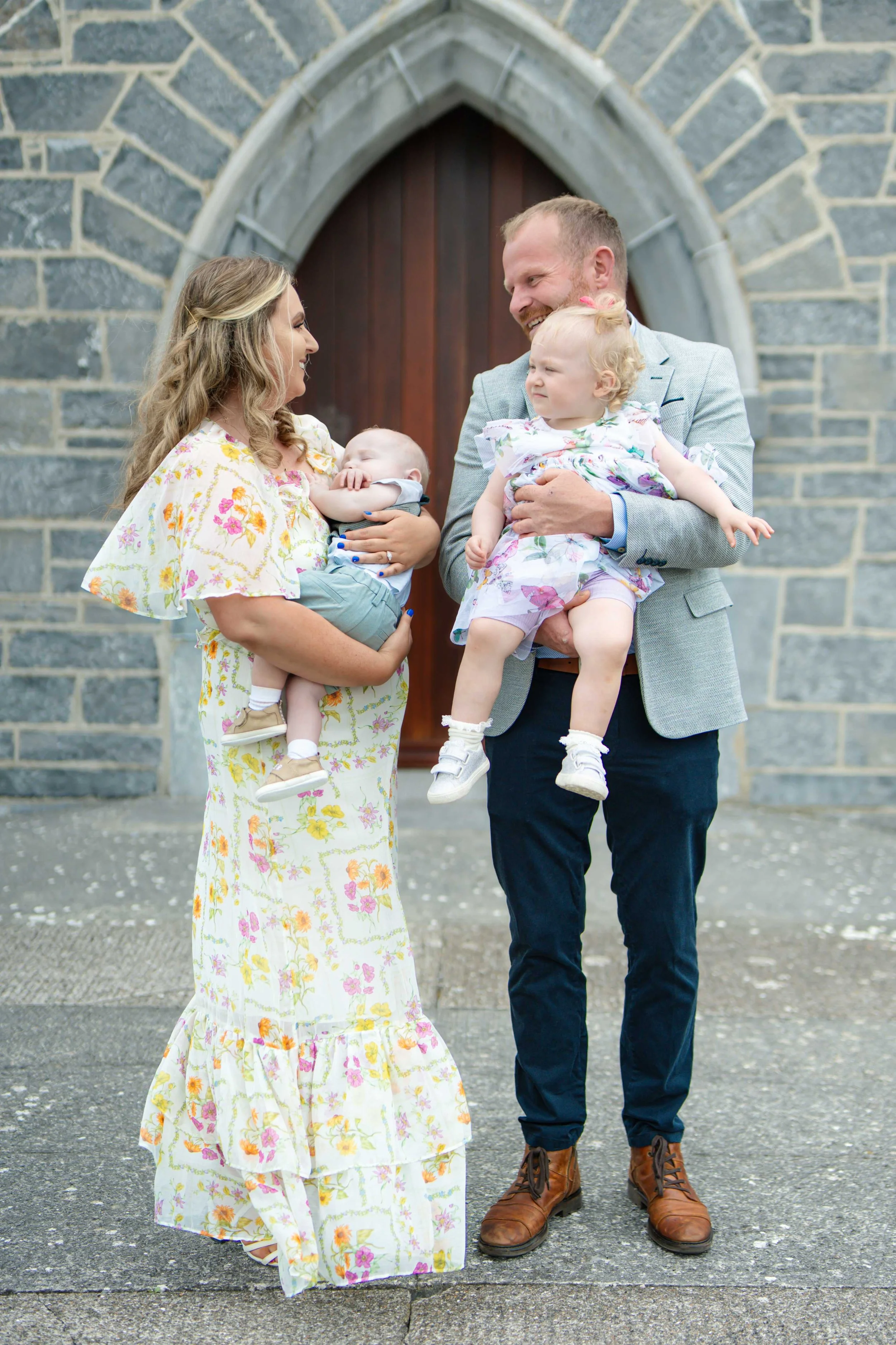 A woman and a man each holding a child, standing outside Ardfert church with a wooden door. They are smiling at each other, and the children are being held close, with the woman’s baby in her arms and the girl in the man's arms.