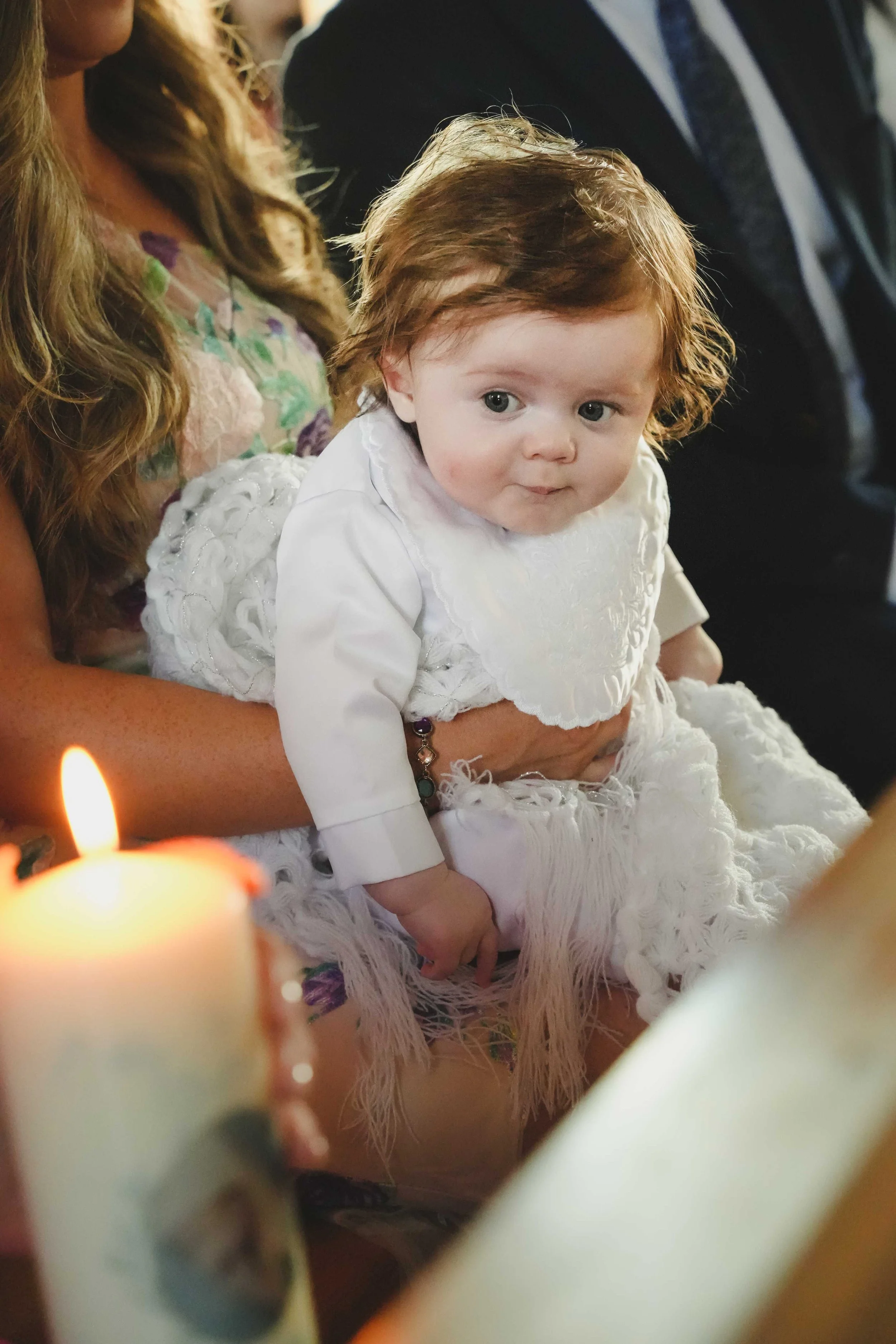 A young child with curly red hair dressed in white, sitting on an adult's lap during christening ceremony in Ballyheigue church surrounded by people, with candles lit in the foreground.