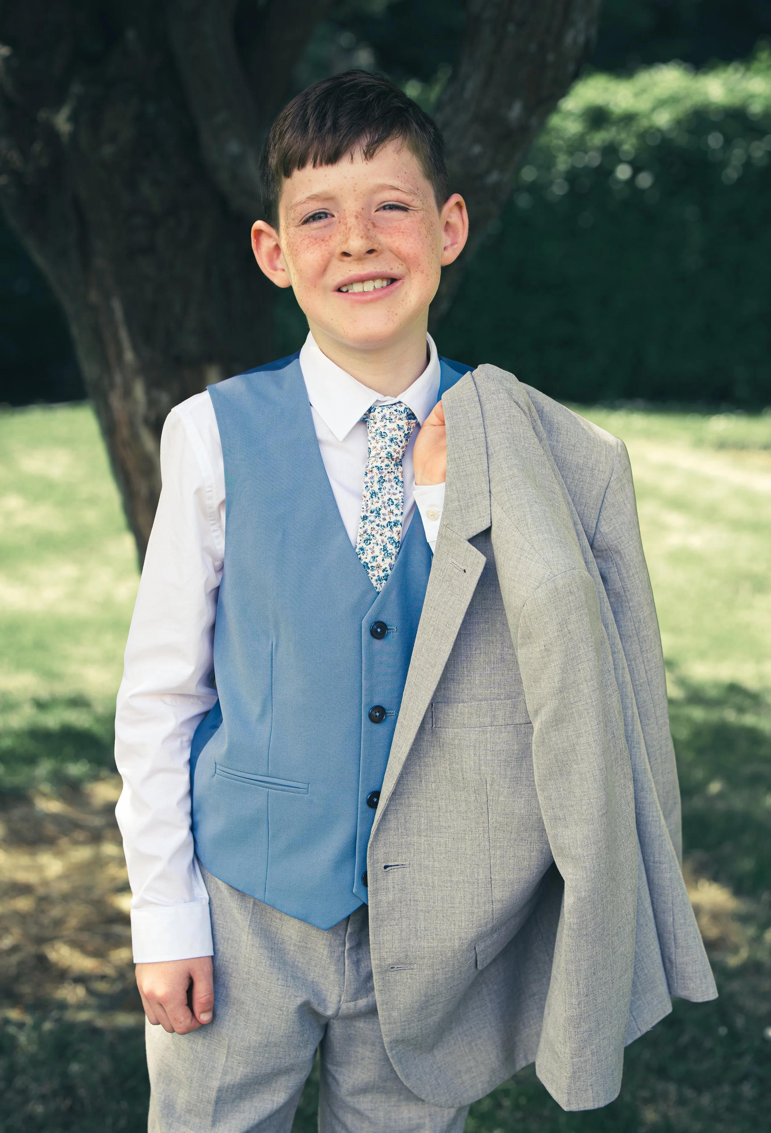 A portrait of a  young boy after his first communion in Town park Tralee, Co.Kerry  holding a gray blazer over his shoulder standing outdoors near a tree with a grassy background.
