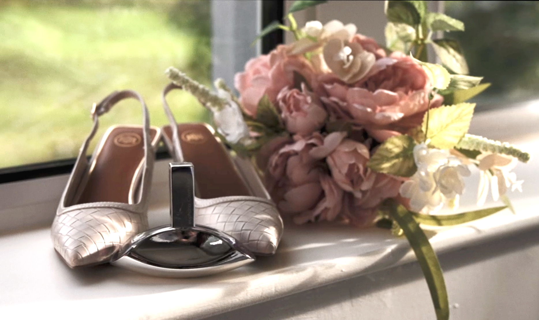A pair of white high-heeled shoes with a woven pattern and elegant design, placed next to a bouquet of pink and white flowers on a windowsill.